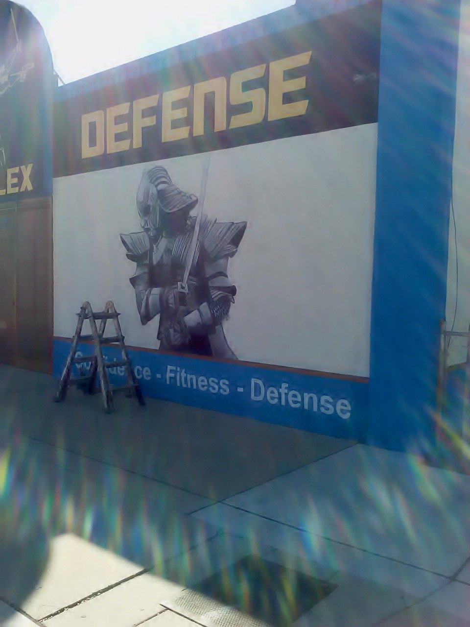 A large promotional wall with the word 'DEFENSE' at the top, featuring an artistic illustration of a person with a helmet and armor reading a book, with a ladder in front of it. The wall also displays the words 'Strength - Fitness - Defense' at the b