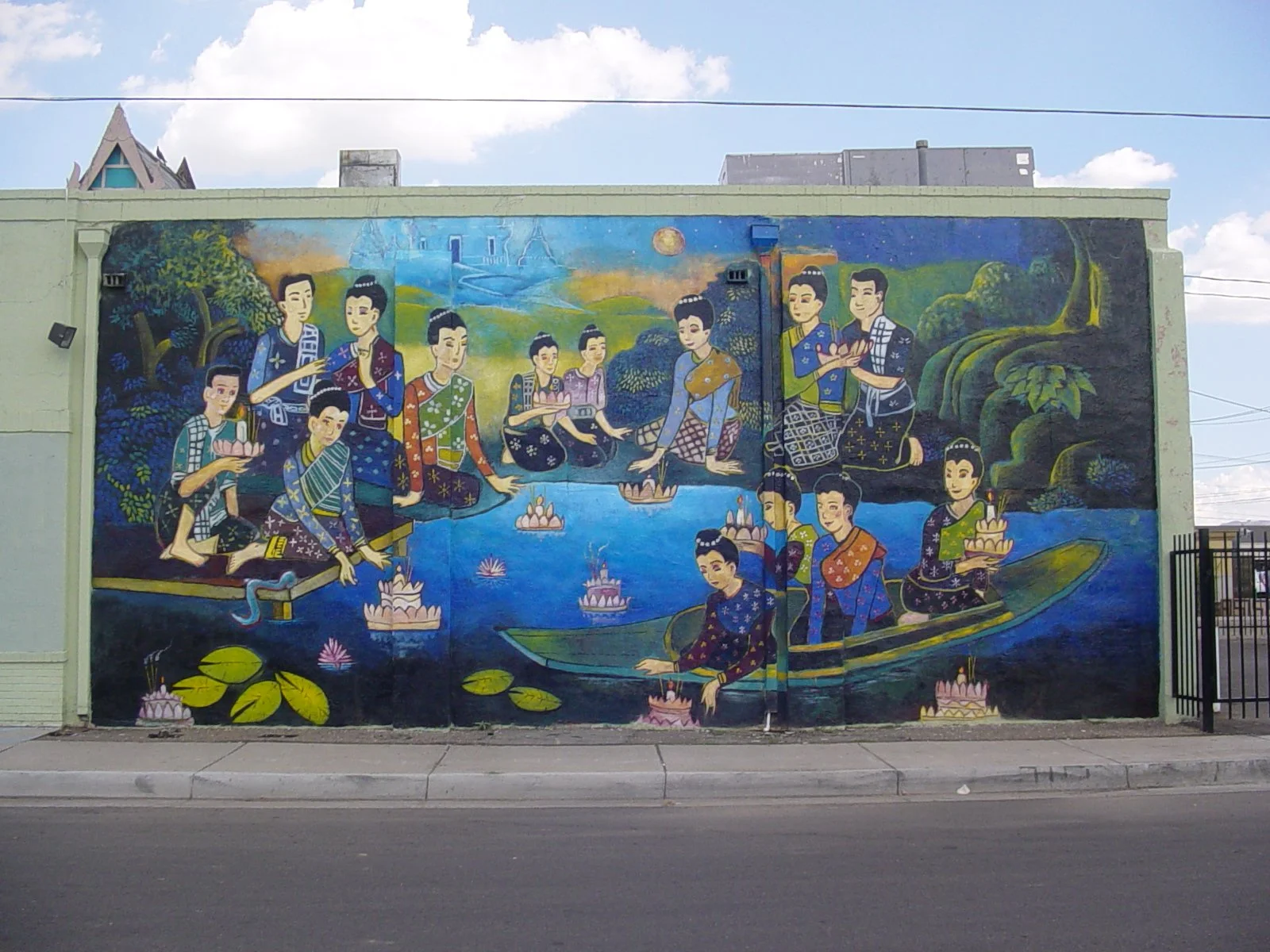 Colorful mural on building wall depicting people in traditional clothing celebrating with candles and lotus flowers by a river, surrounded by greenery and trees.