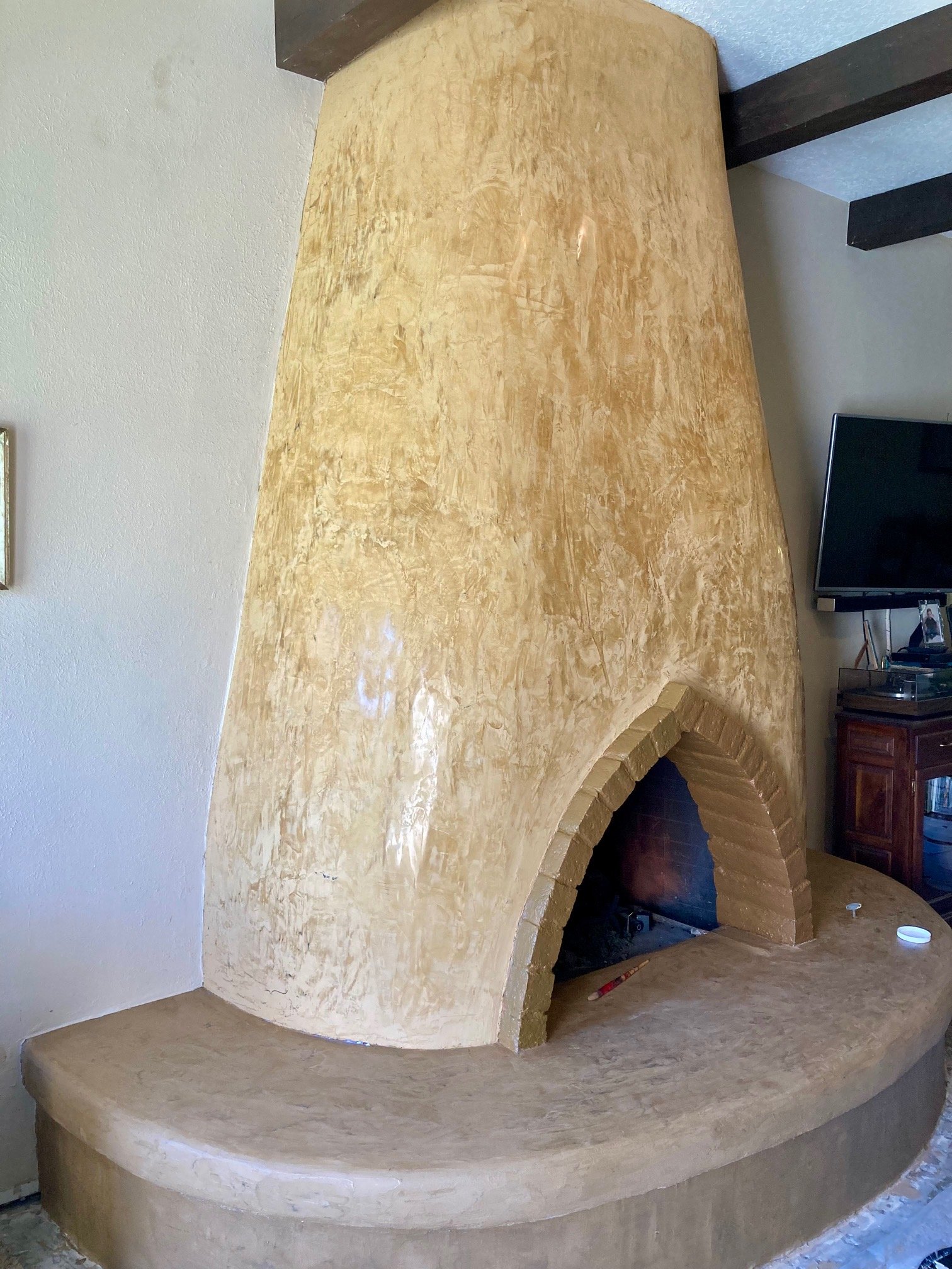 A large, beige stucco fireplace with an arched opening in a living room, with a TV and wooden cabinet to the right.