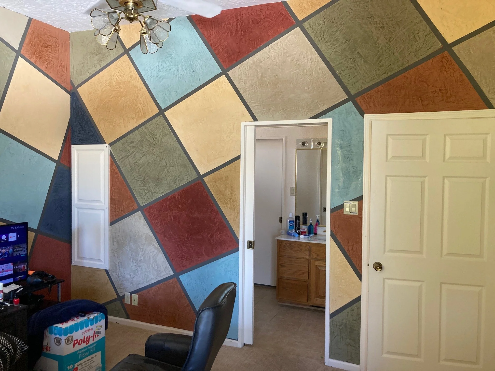 A room with a colorful geometric patterned wall, a ceiling fan, a computer desk with a monitor, a black office chair, and an open doorway leading to a bathroom with a vanity and toiletries.