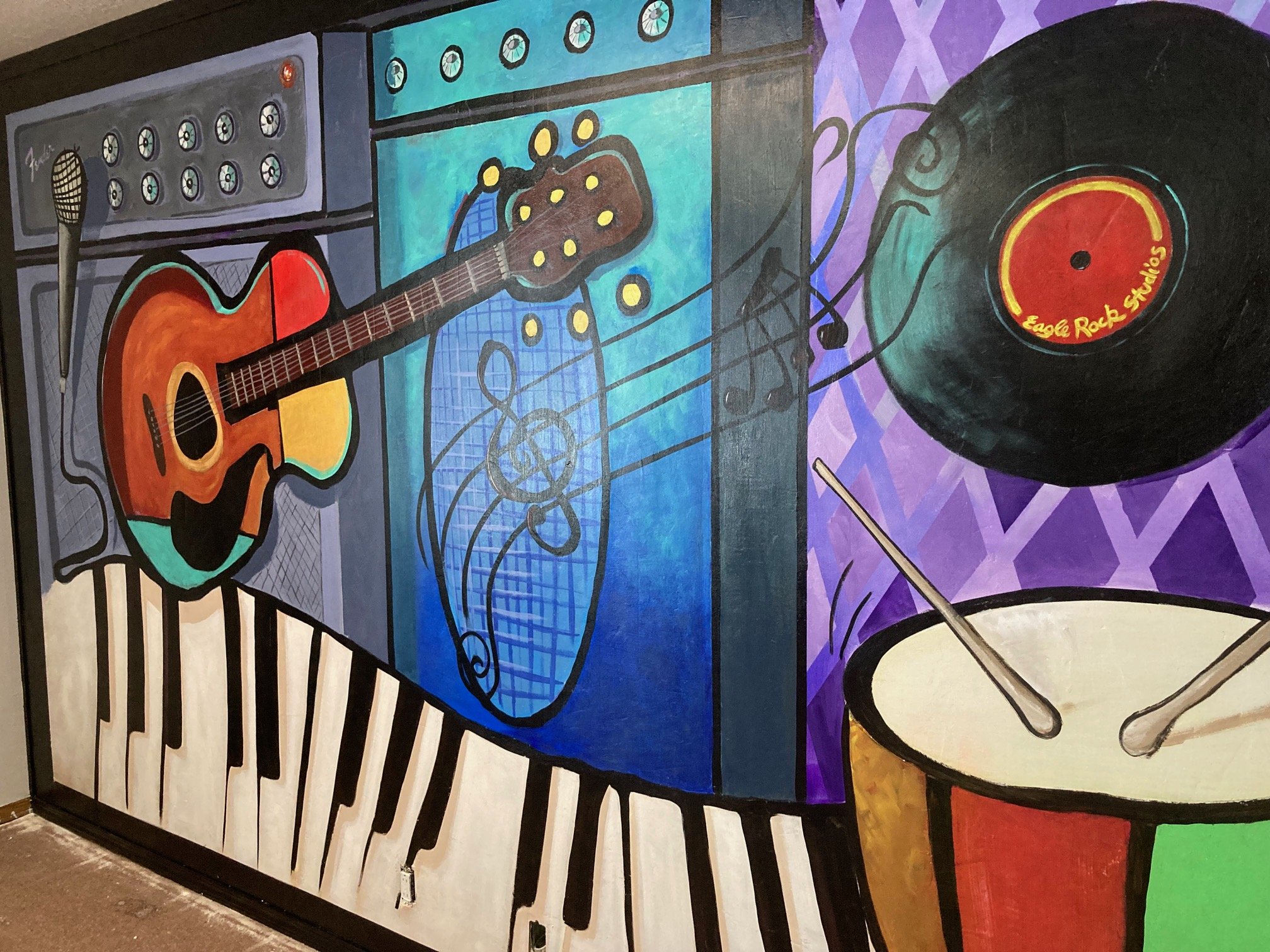 Colorful mural featuring musical instruments: an acoustic guitar, a microphone, a vinyl record labeled "Eagle Rock Studios," a drum with drumsticks, and piano keys. The background includes amplifiers and a staff with musical notes.