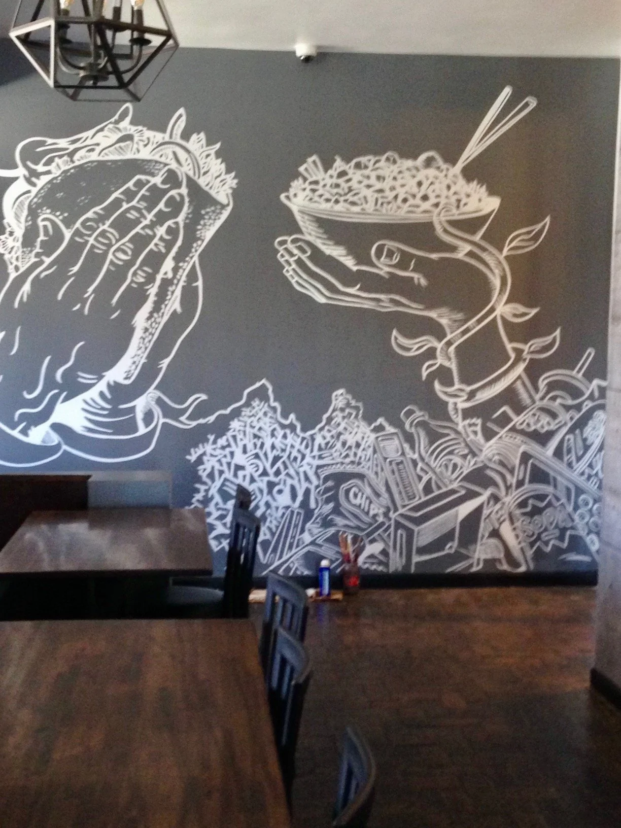 Chalkboard mural in a restaurant showing a hand holding a burger, a hand holding a bowl of noodles, and a pile of trash with bottles, boxes, and straws.