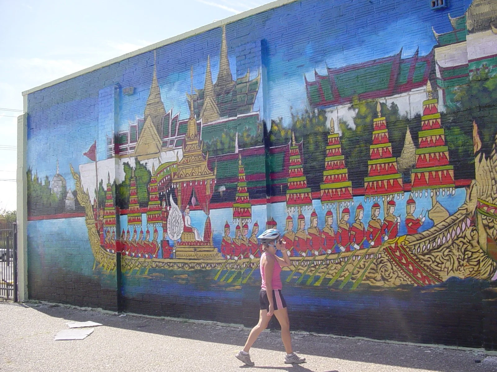 Mural painting of a traditional Thai royal barge on a blue water background, with a woman wearing a helmet and pink tank top walking in front of it.  Custom mural and art work for a business, Billings, Montana.