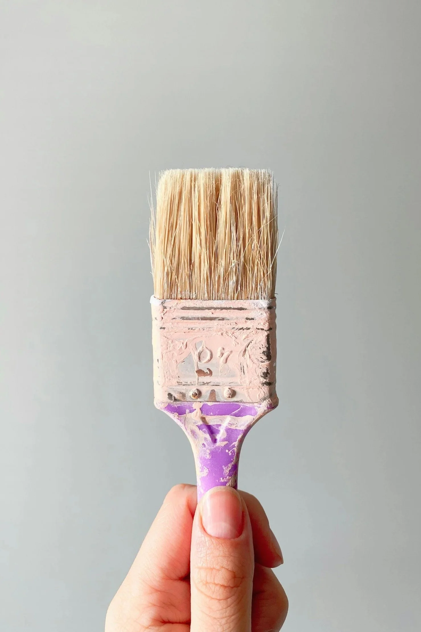 Close-up of a paintbrush with light pink paint on bristles, held by a person's hand against a plain, gray background.