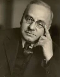 A black and white portrait of a man wearing glasses, with one hand resting on his face, looking thoughtfully at the camera.