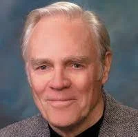 Portrait of an older man with gray hair wearing a gray blazer and a dark shirt, smiling against a blue background.
