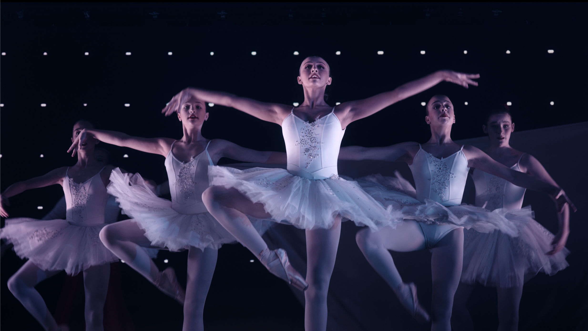 Ballet dancers performing on stage in white tutus, with stage lights overhead.