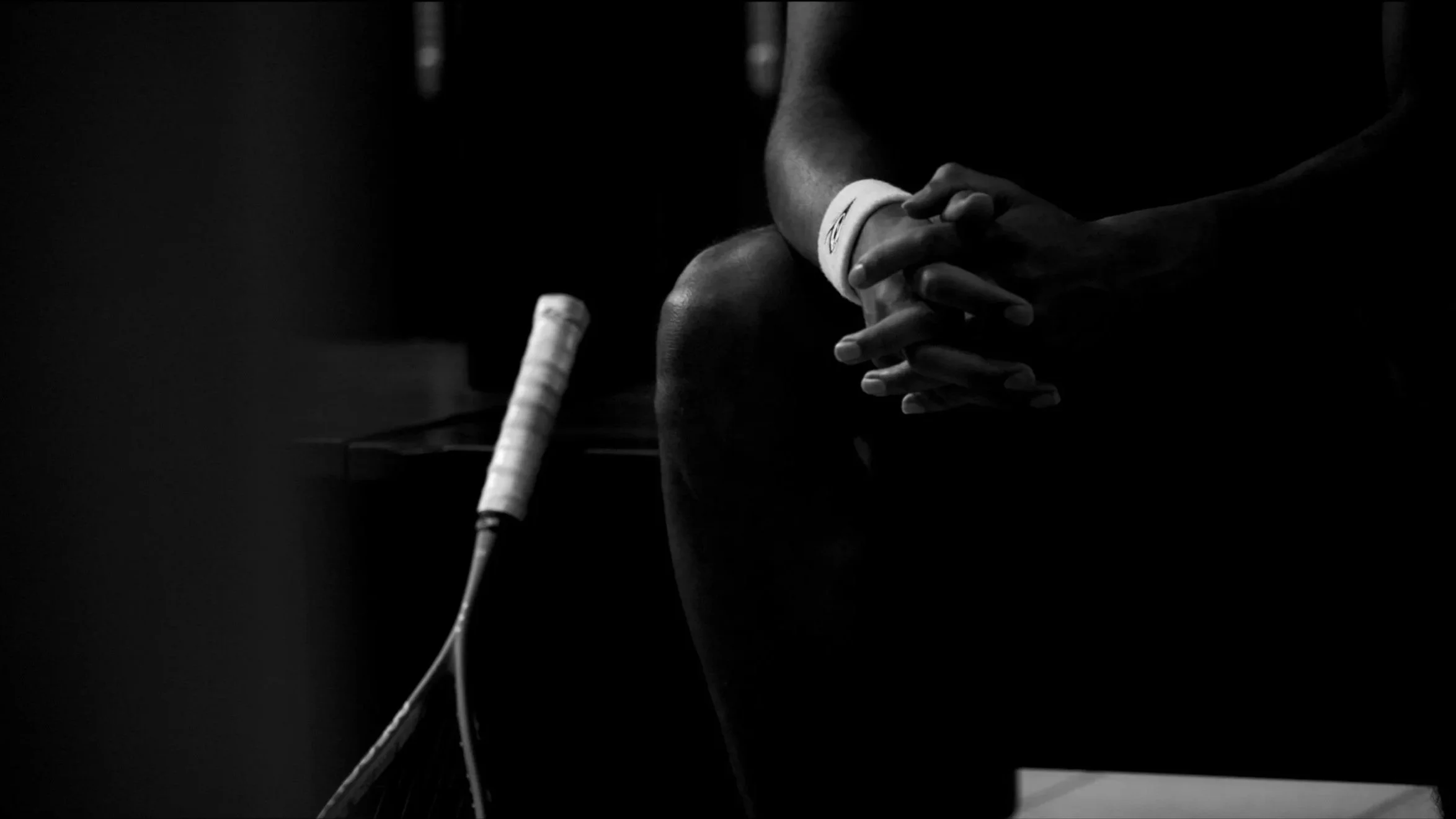 A person sitting with hands clasped, wearing a wristband, near a tennis racket on a table, in a dark setting.