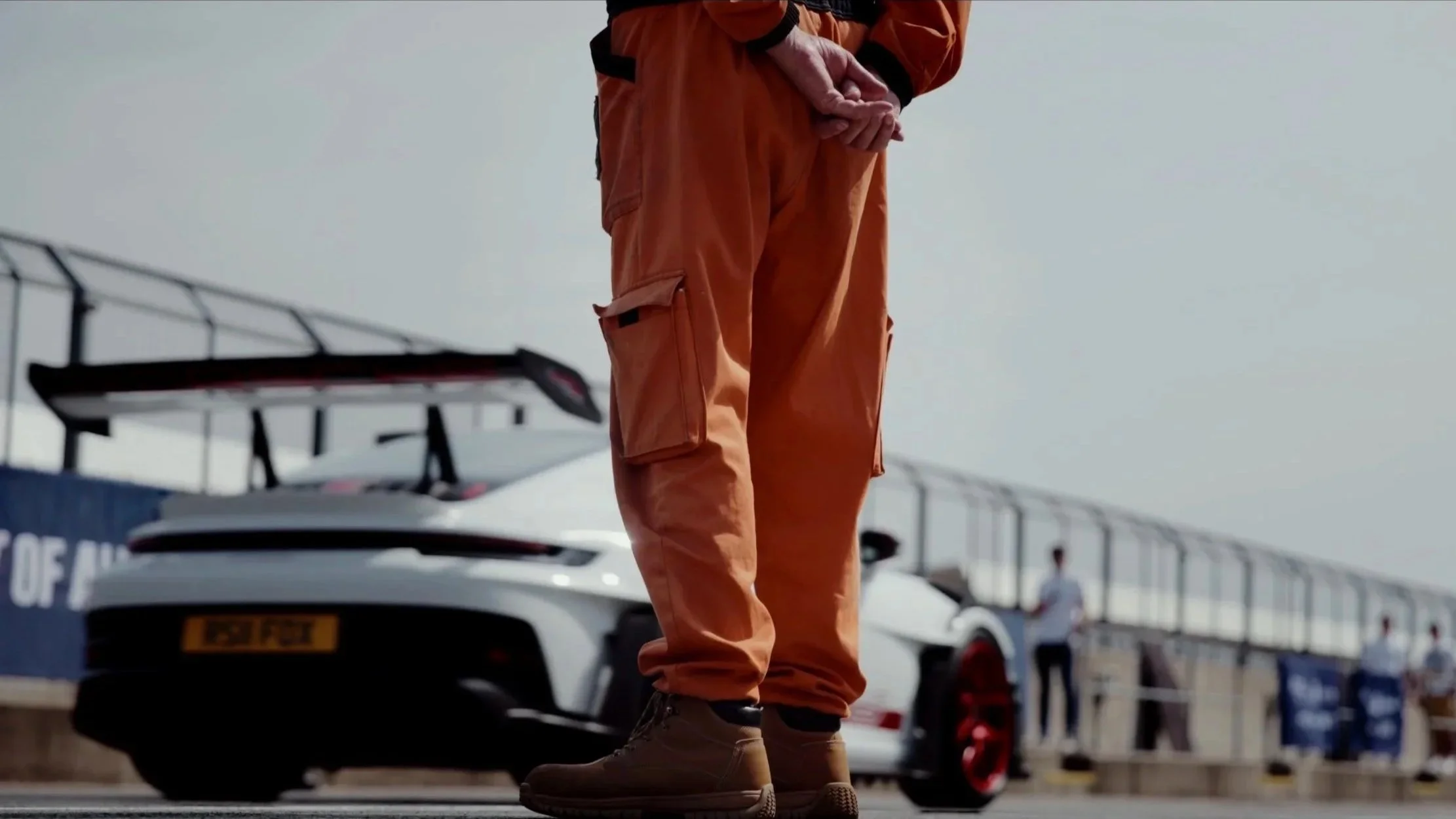 Person standing in front of a racing car at a race track, partially visible with orange racing suit and brown boots.