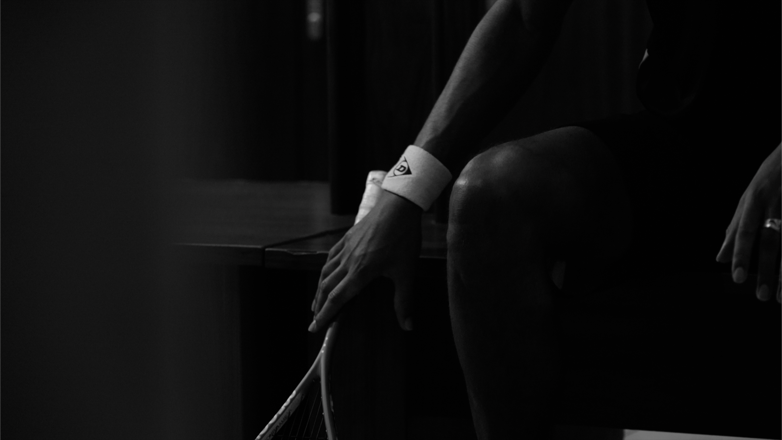 A person sitting on a bench, holding a tennis racket, wearing a wristband, in a dark, low-light setting.