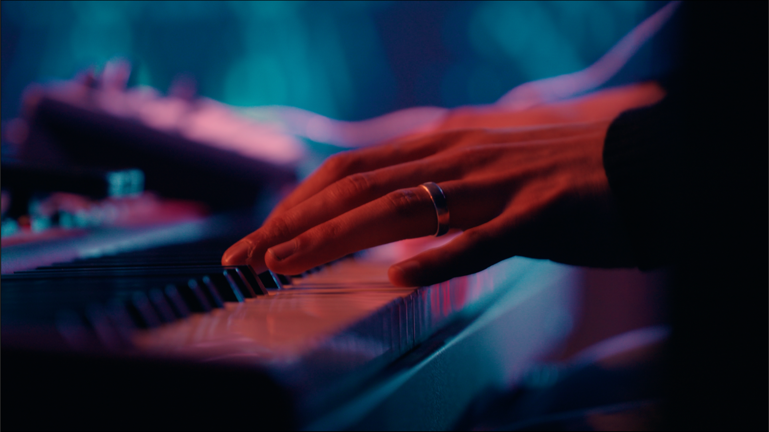 Close-up of a person's hand with a ring playing a piano keyboard in dim, colorful lighting.