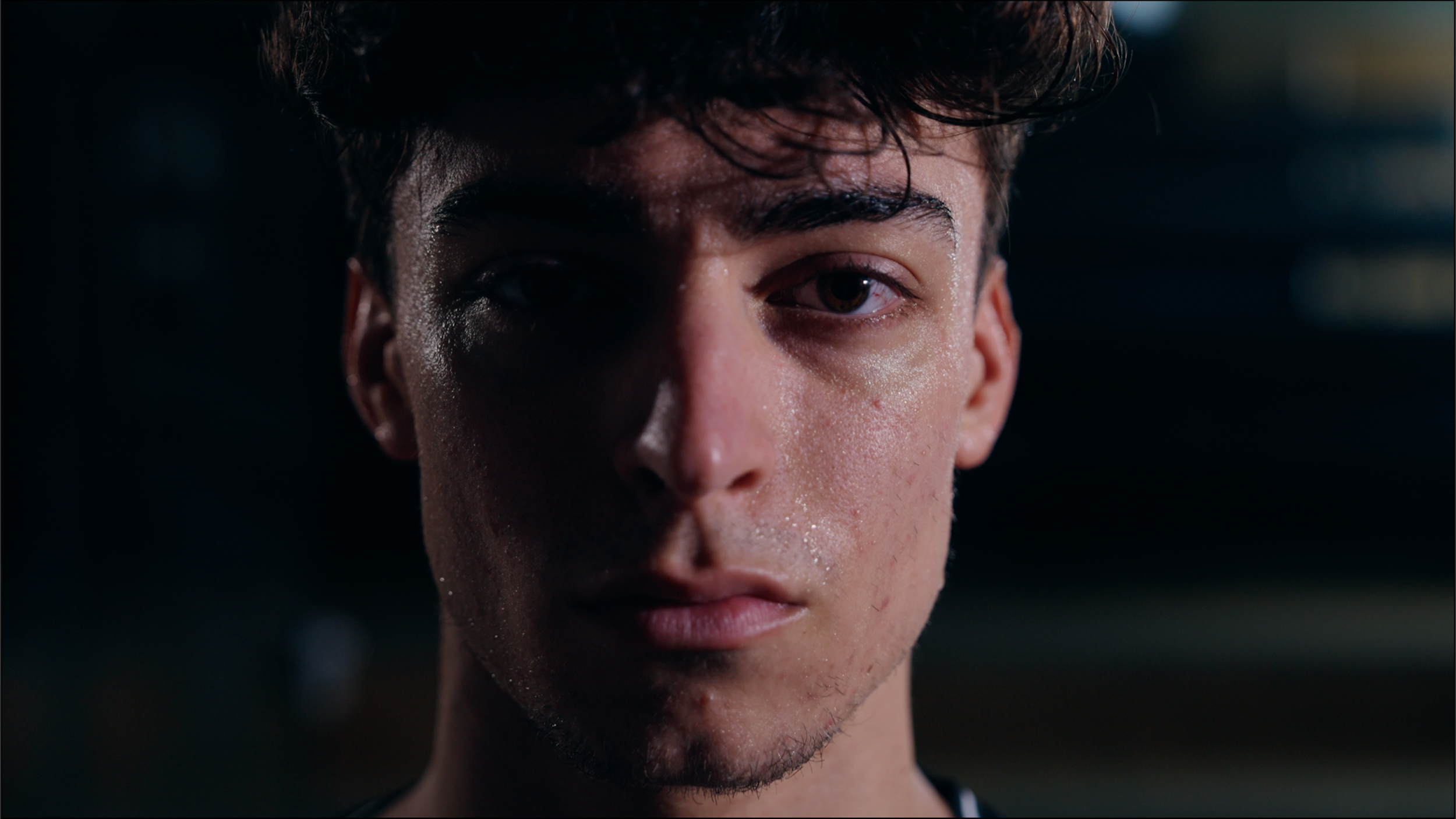 Close-up of a young man's face with sweat, dark background, serious expression.