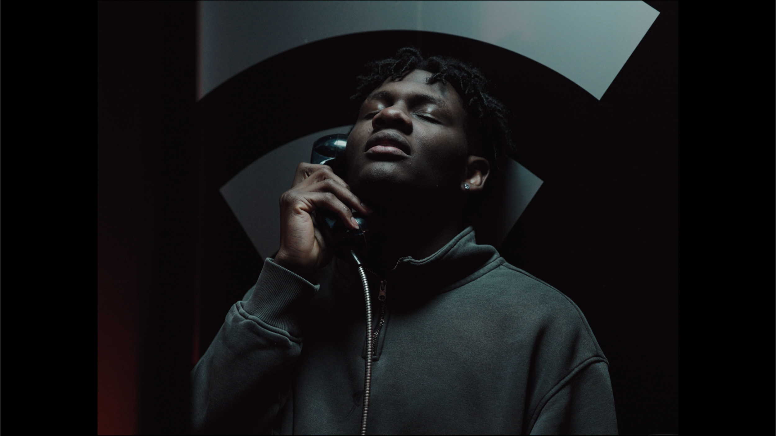 A young man with dark skin and dreadlocks is holding a vintage phone receiver to his ear with his eyes closed in a dark room.