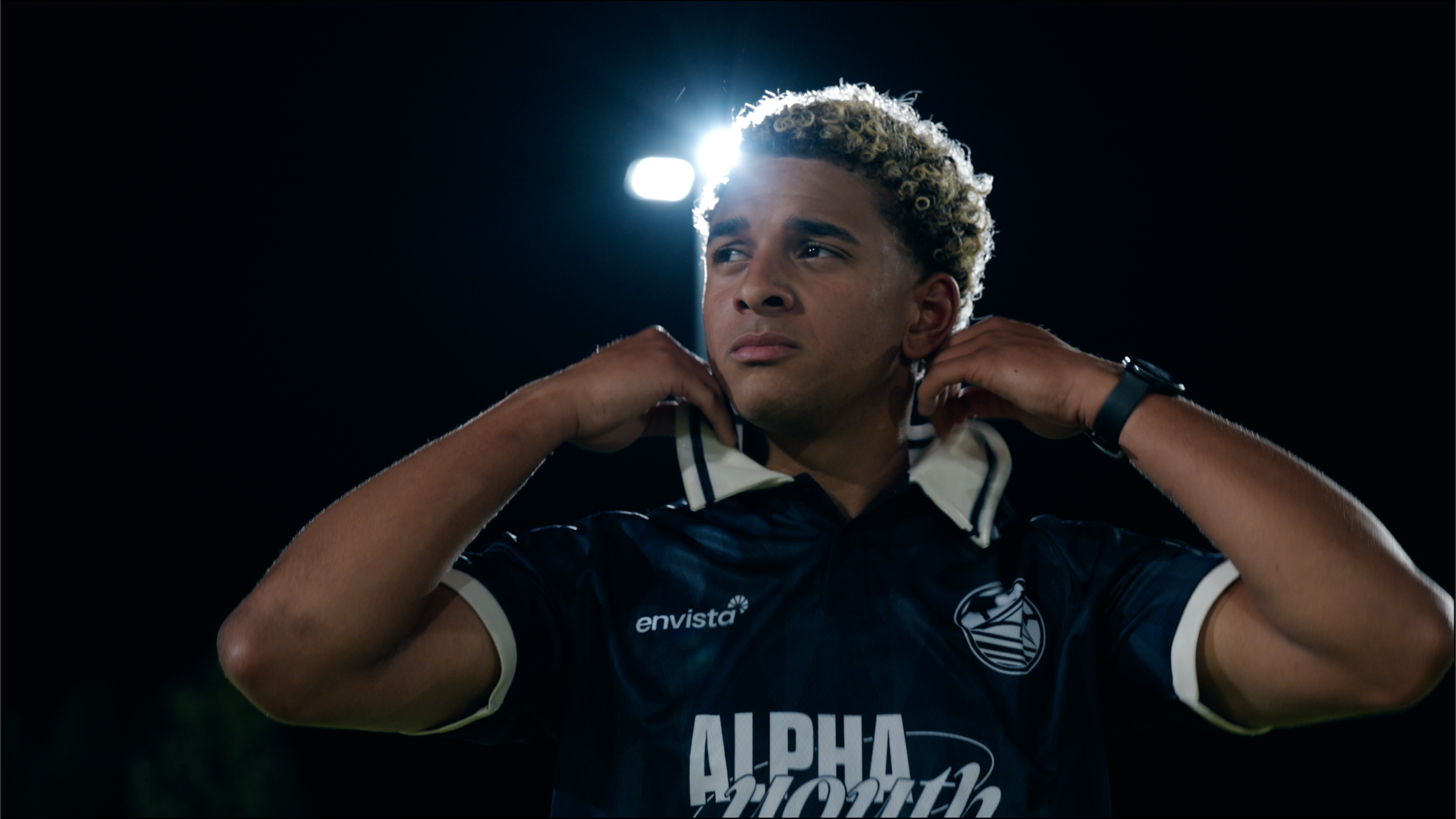 A young man with curly blonde hair adjusting his sports jersey under bright stadium lights at night.