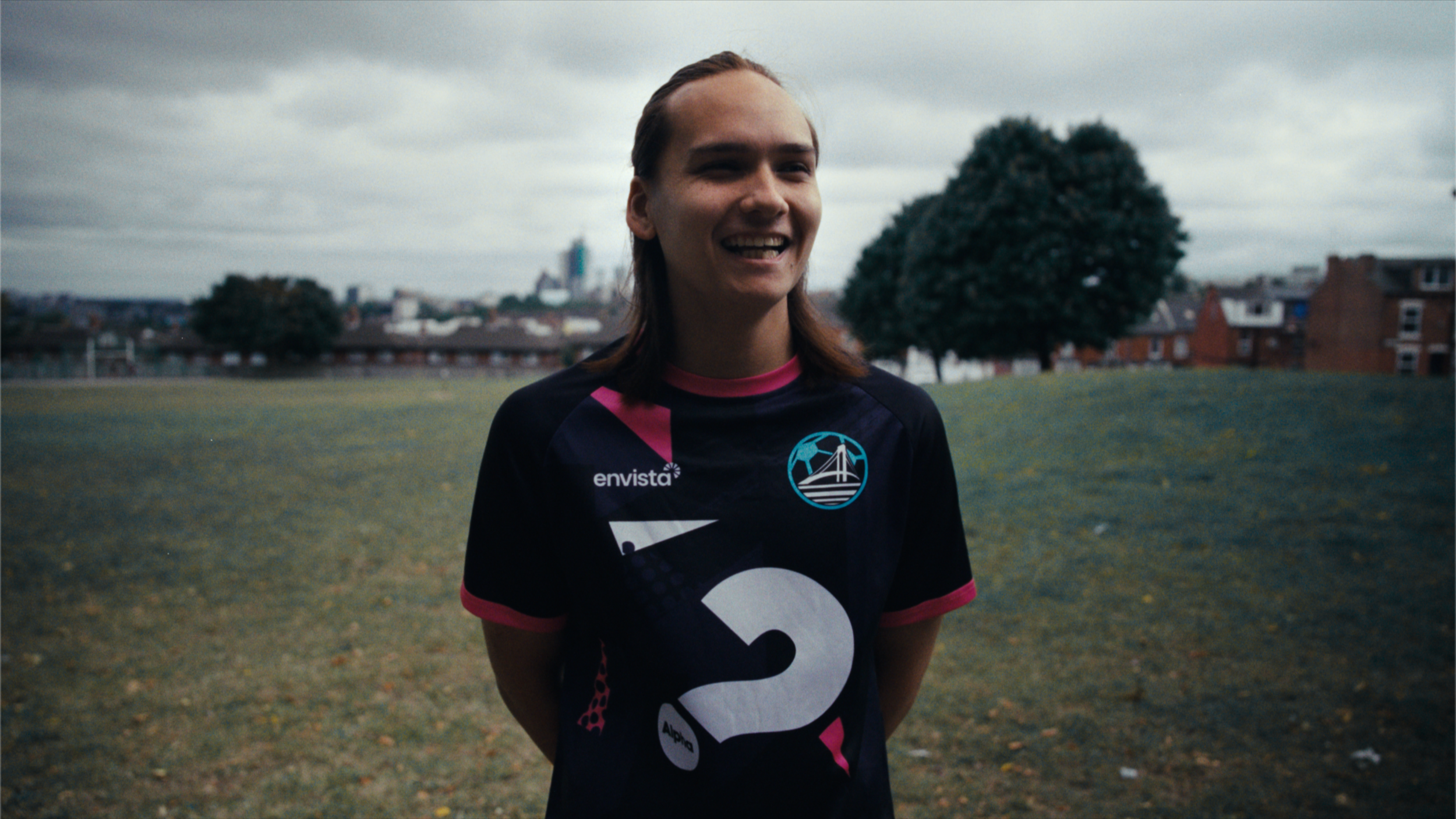 Young woman in a black sports jersey standing outdoors on a cloudy day, smiling with trees and buildings in the background.