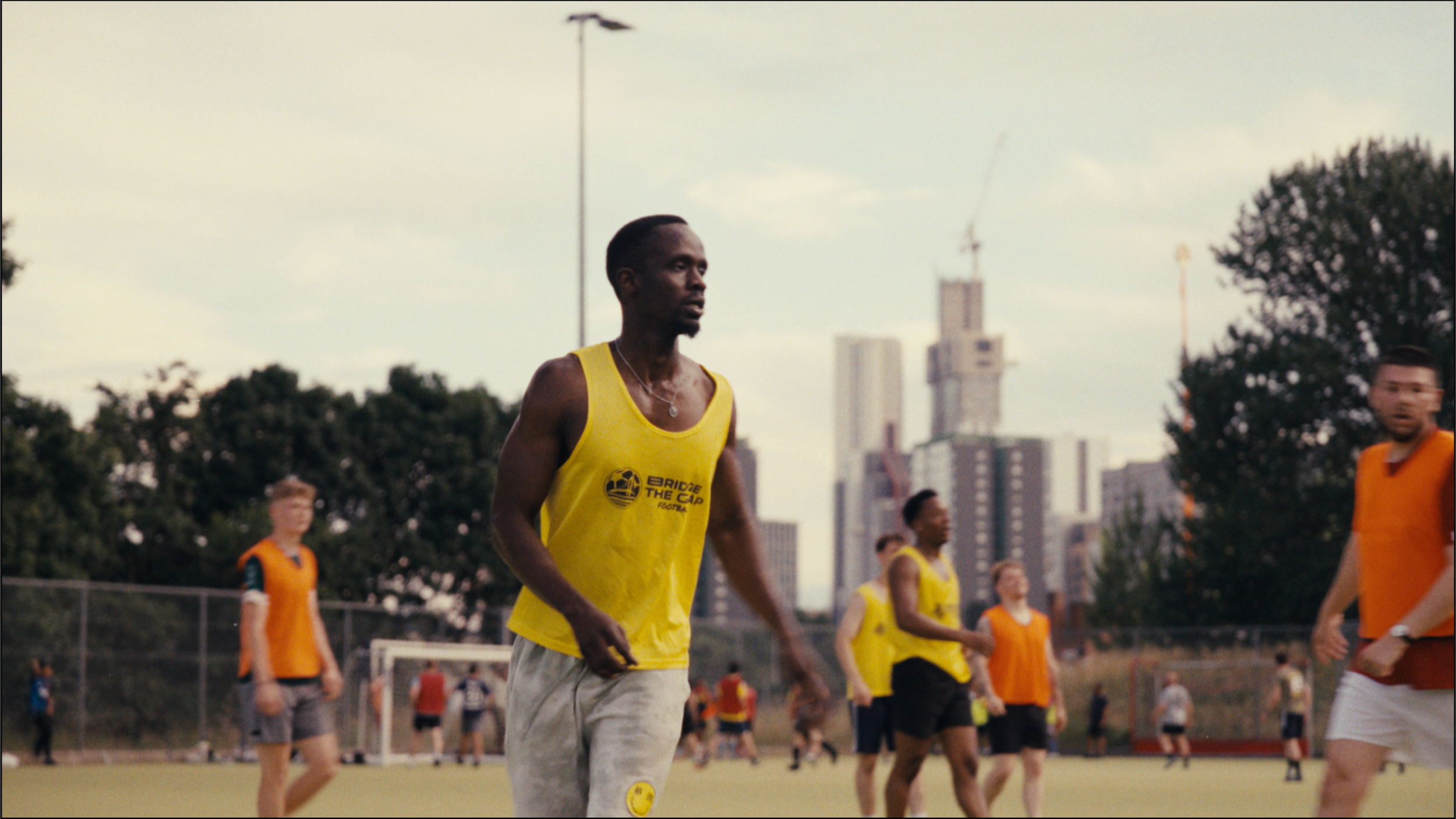 A group of people playing soccer on a field, with some wearing yellow and orange jerseys, with a city skyline in the background.