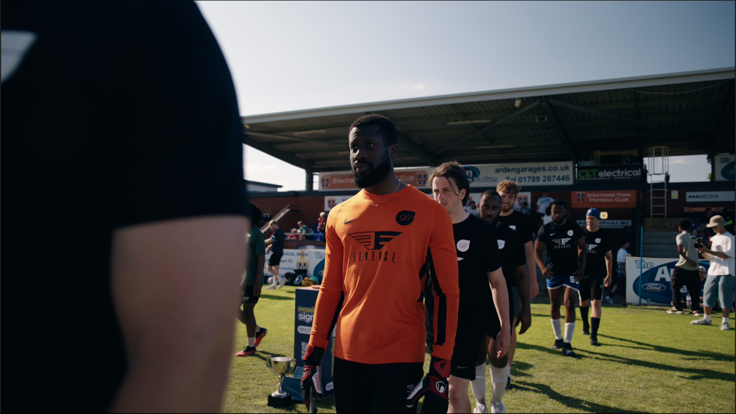 Soccer players walking onto the field before a match, with a goalkeeper in orange leading, against a backdrop of a small stadium and spectators.