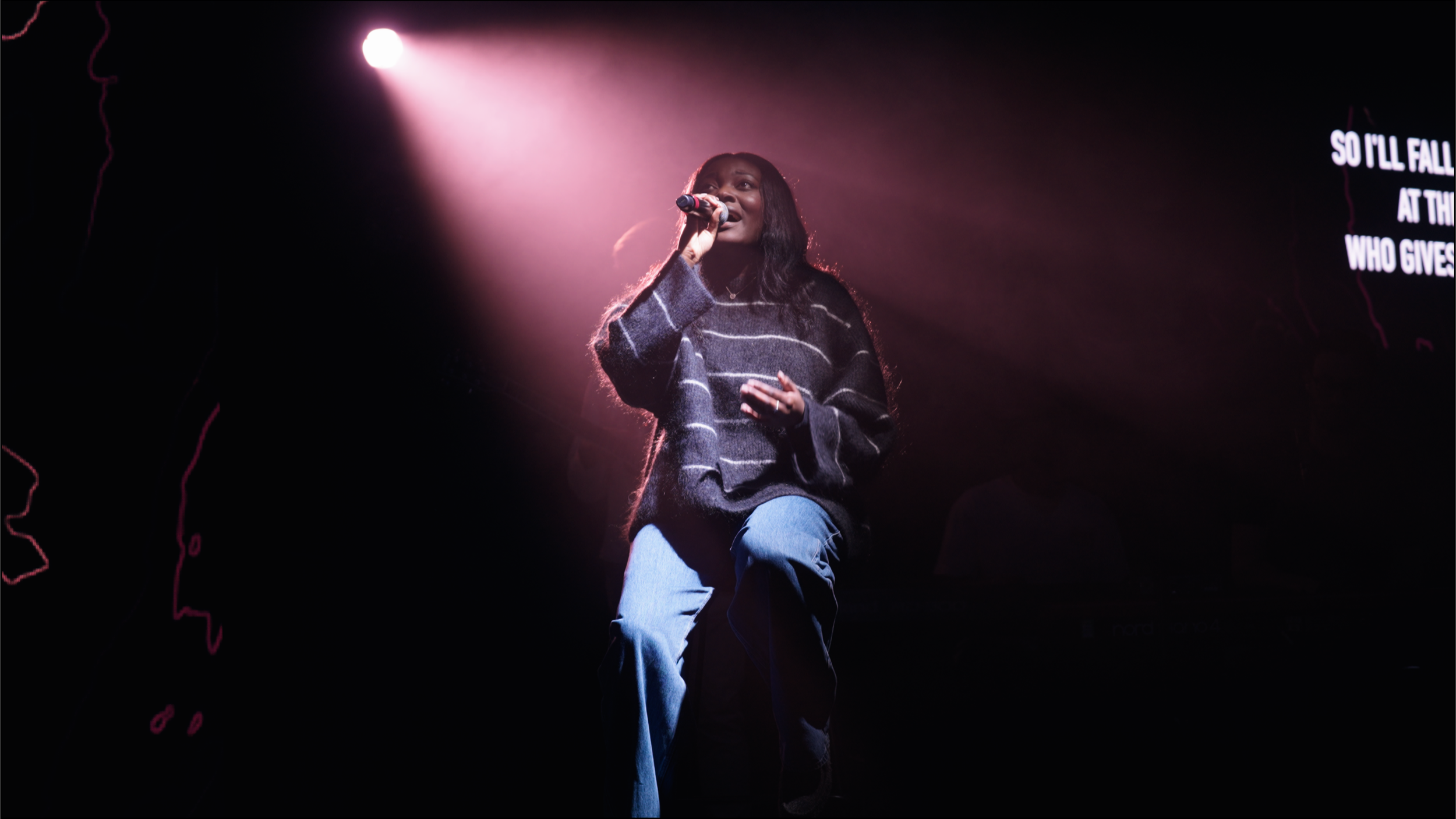 A woman singing into a microphone on stage, illuminated by pink spotlights, with partial lyrics displayed on a screen behind her.