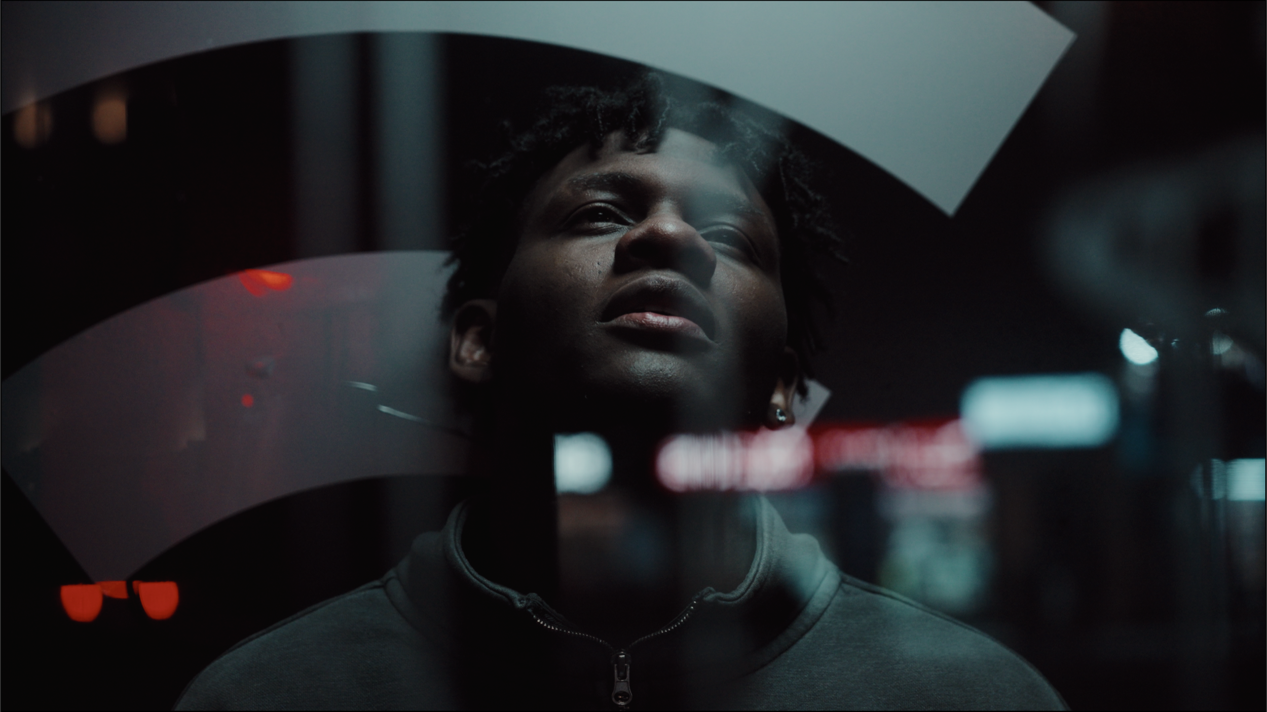 A person with dark skin and short curly hair looking up through a glass window with reflections of city lights at night.