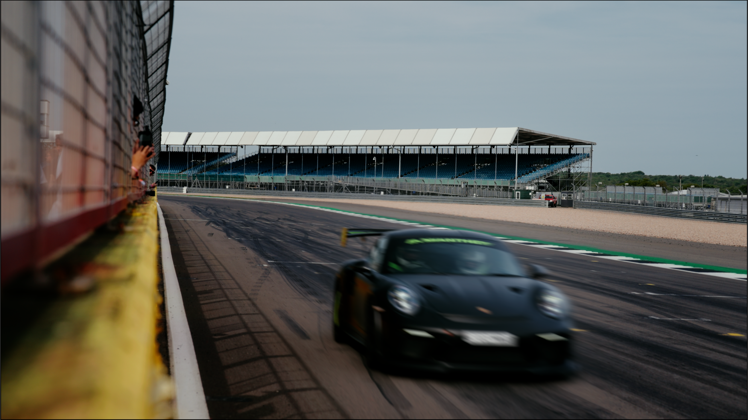 A black race car speeding on a race track with a blurred background of grandstands and a right-side tire wall.