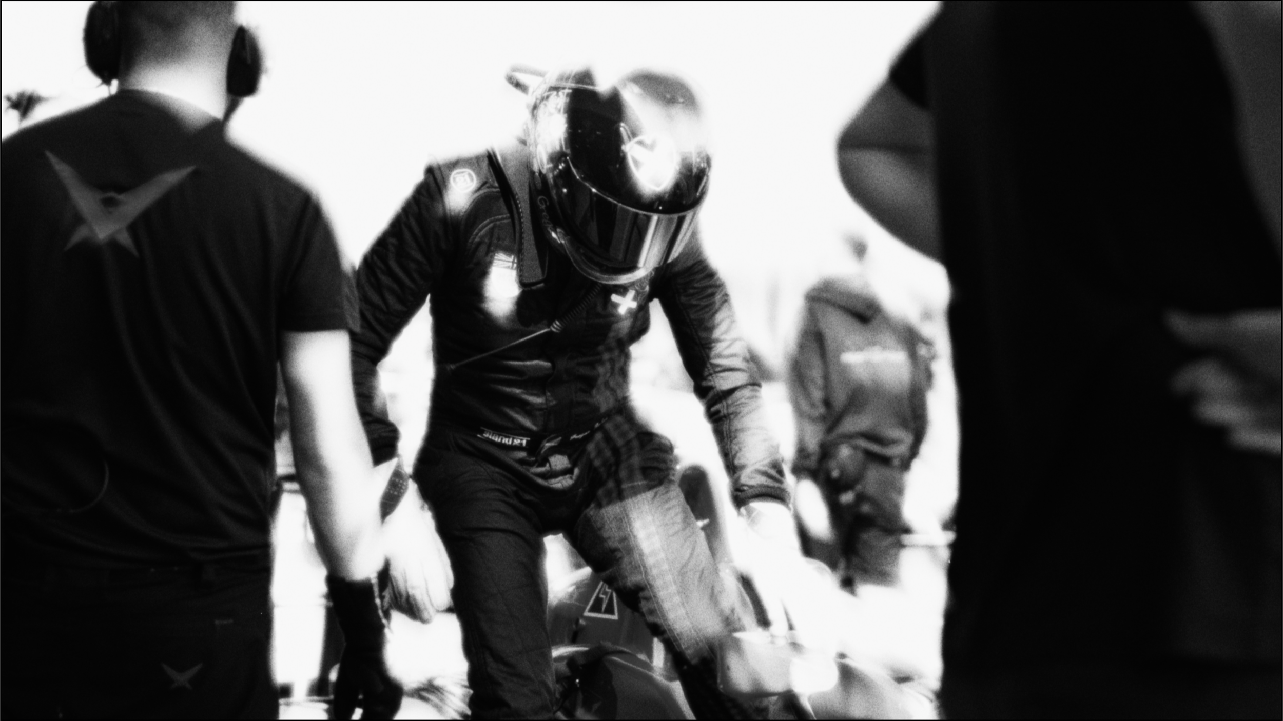 A group of racers preparing for a race, with one central figure wearing a helmet and racing suit getting on a motorcycle, while others stand nearby.