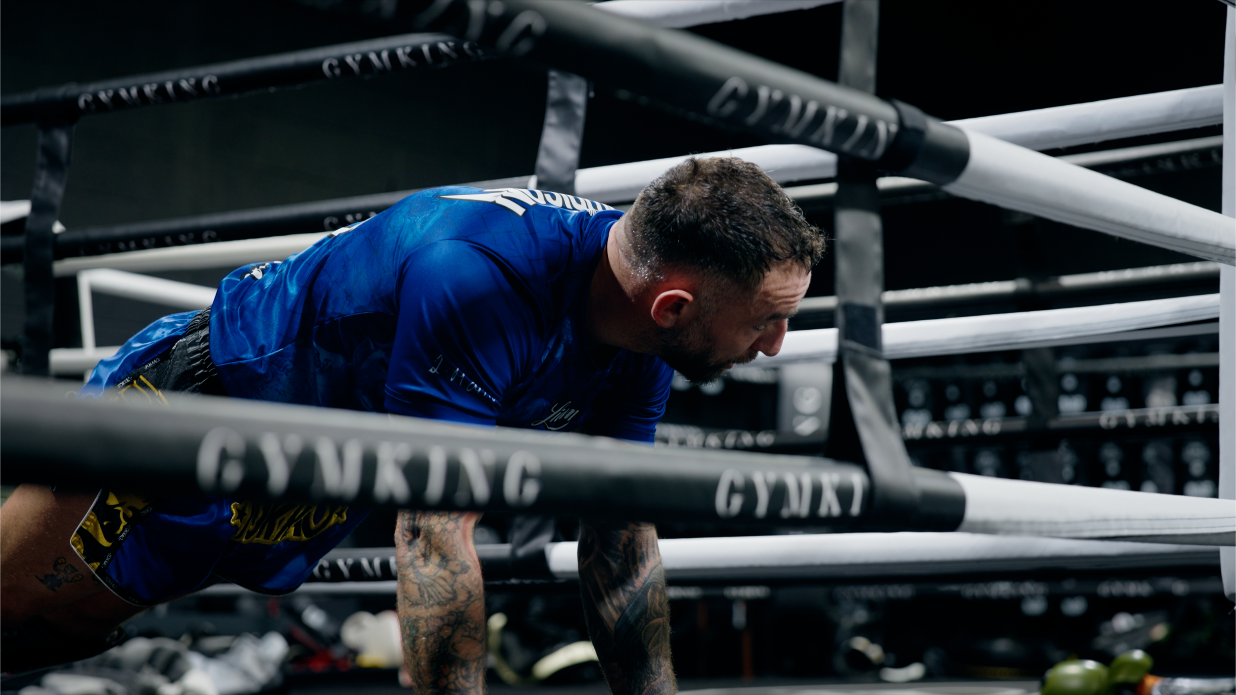 A man with tattoos on his arms doing push-ups in a boxing gym. He is wearing a blue shirt and black shorts, surrounded by boxing ring ropes and gym equipment.