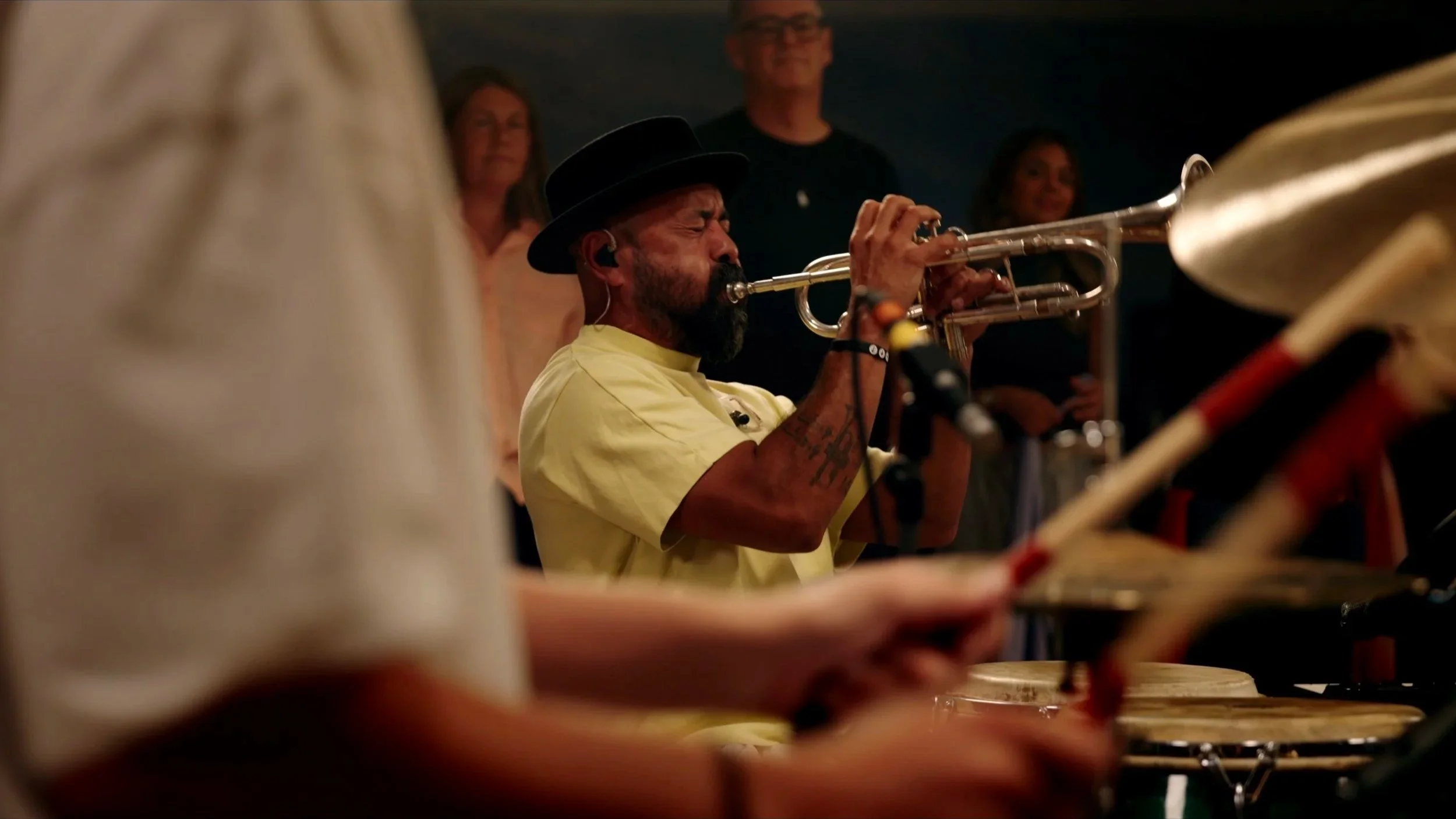 A man with a beard and hat playing the trumpet during a musical performance, with a few people in the background watching.