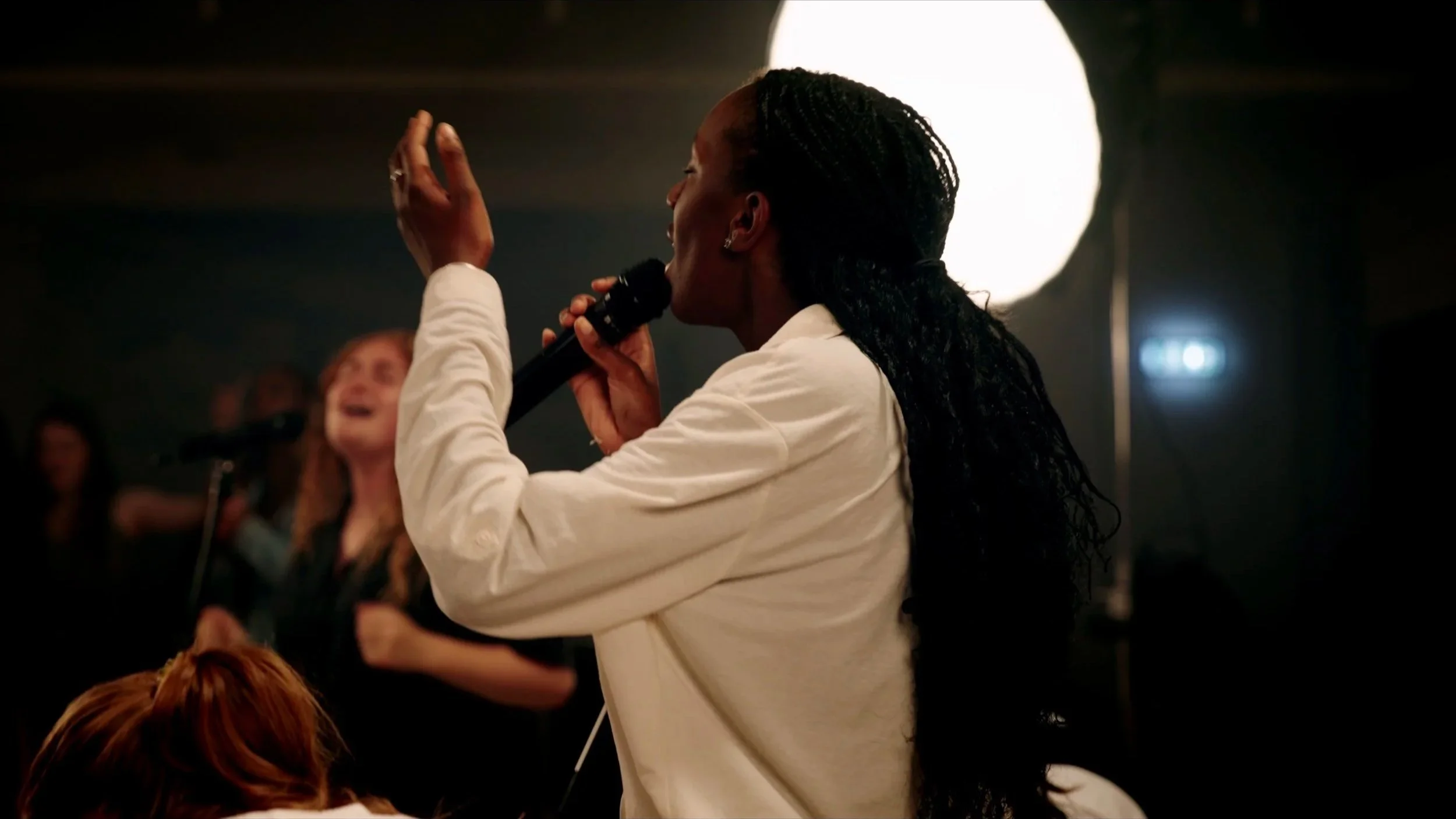 A woman with long braided hair singing into a microphone on stage, with people in the background.