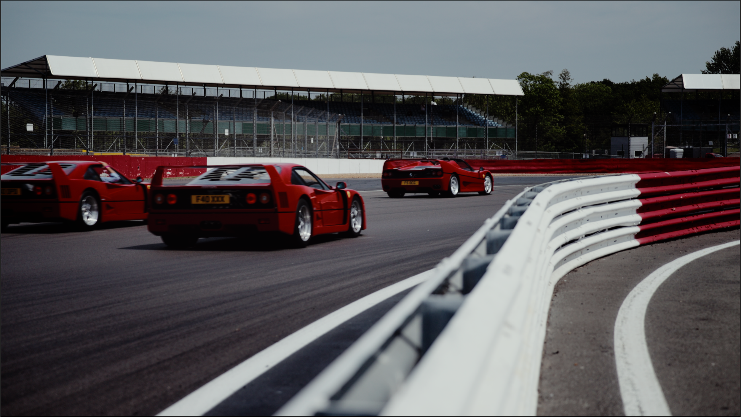 Three red sports cars racing on a track, with a white and red barrier on the side and grandstands in the background.
