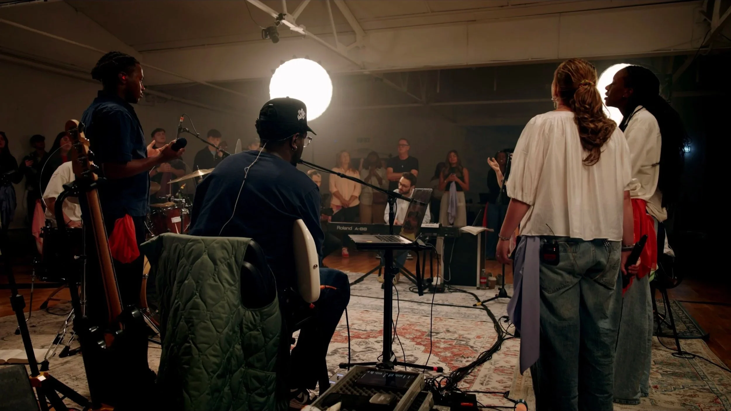 Musicians and singers perform in a studio with musical instruments, a keyboard, and an audience watching.