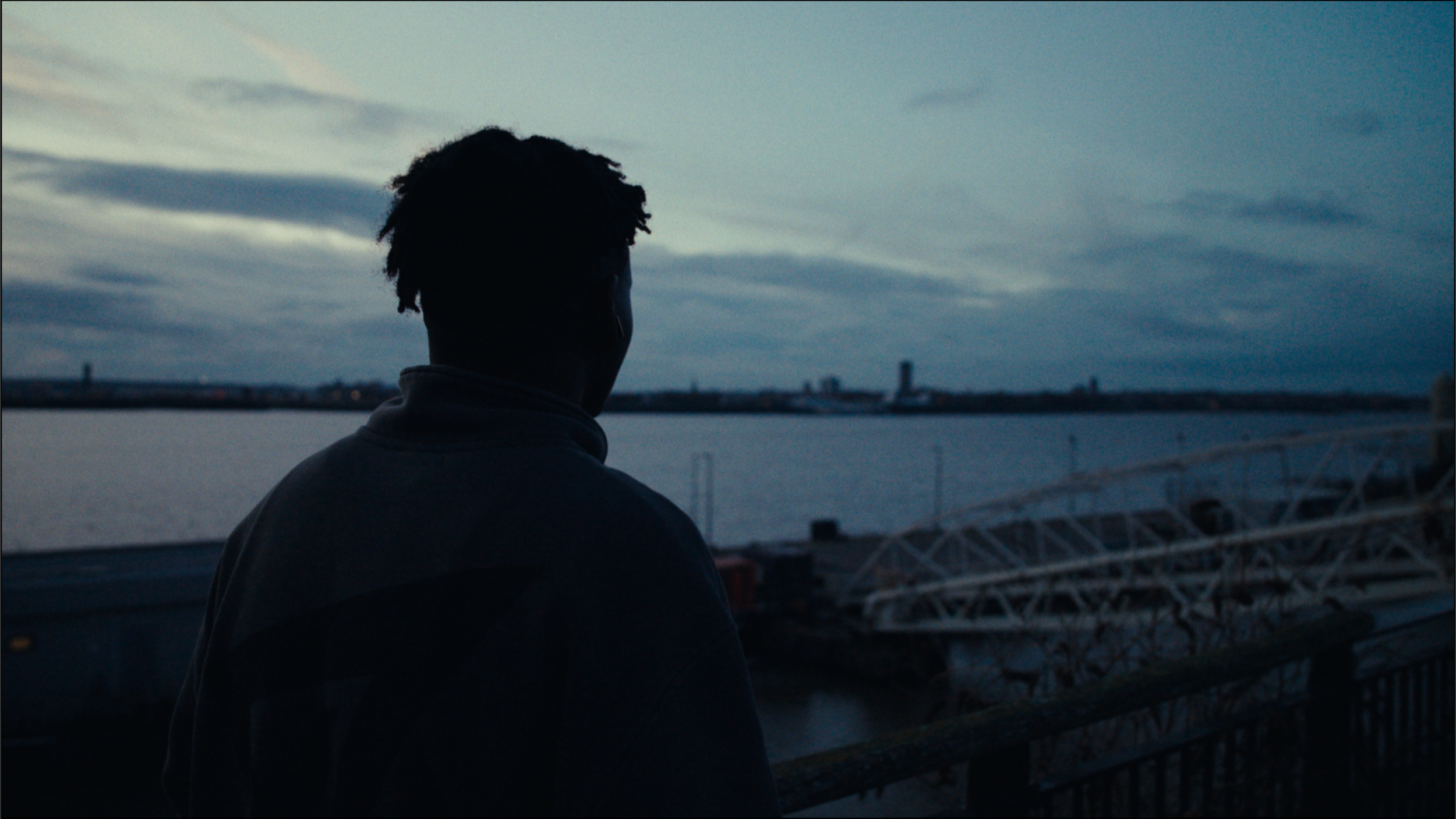 A person with short curly hair stands on a waterfront at dusk, looking out over the water with a city skyline in the distance under a cloudy sky.