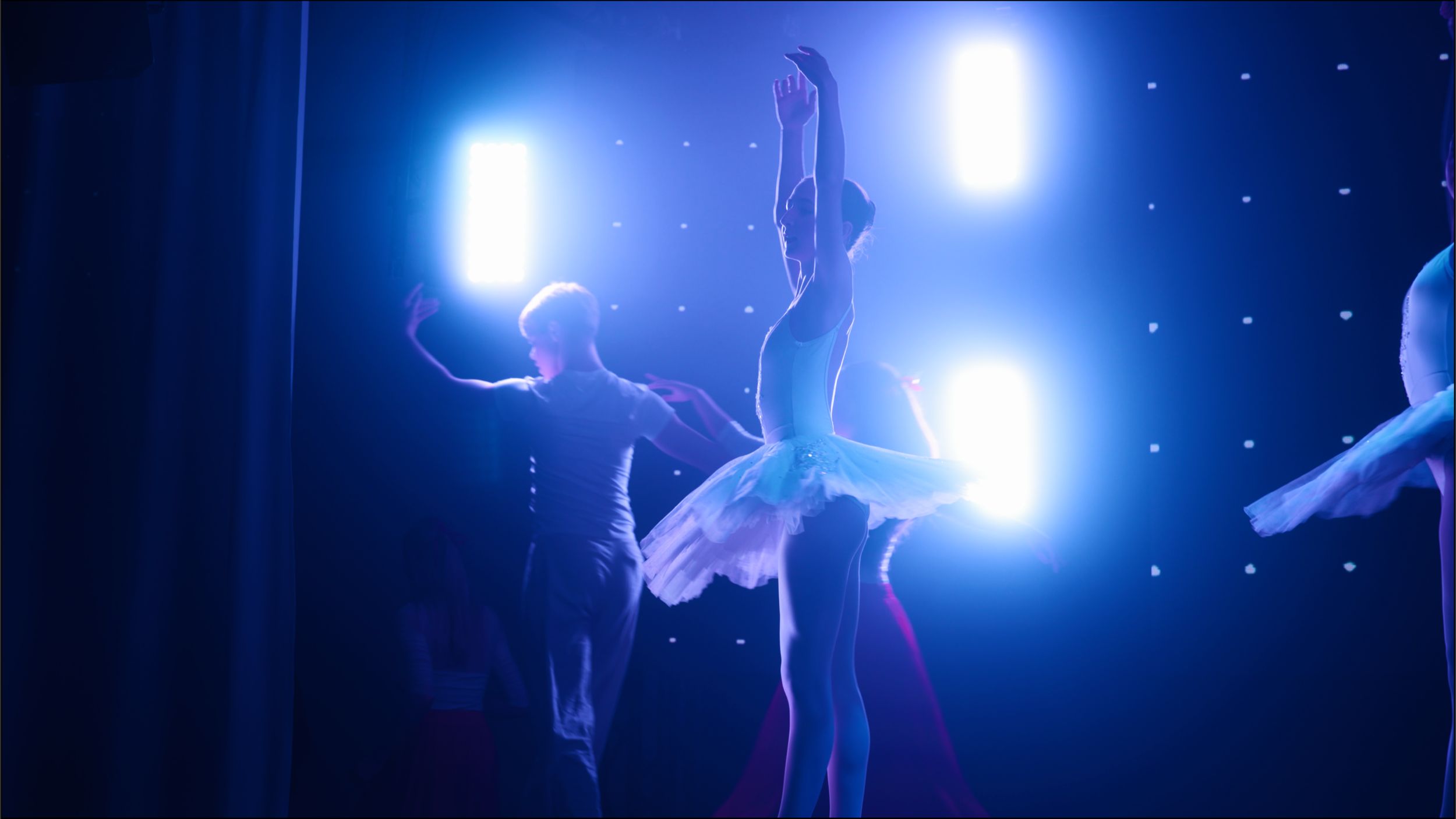 Ballet dancers on stage under bright blue lights, some with arms raised and wearing tutu costumes.