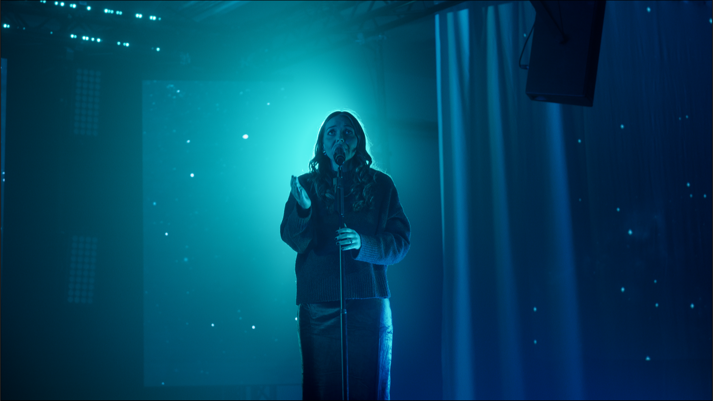 A woman singing into a microphone on a dimly lit stage with blue lighting and starry background.