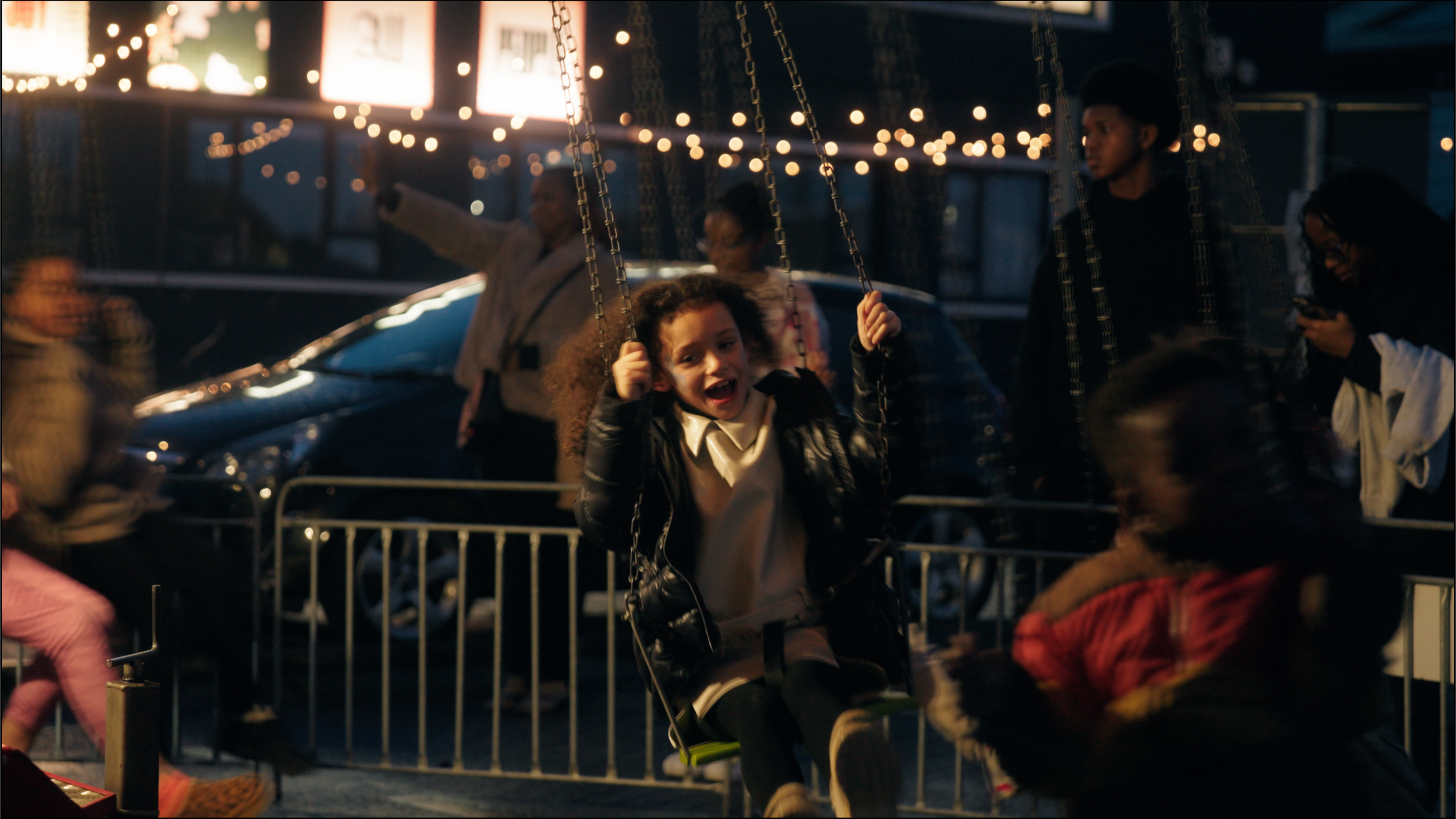A young girl with curly hair wearing a black jacket and white shirt is smiling while swinging on a swing at a carnival during the evening, with other people and illuminated signs in the background.
