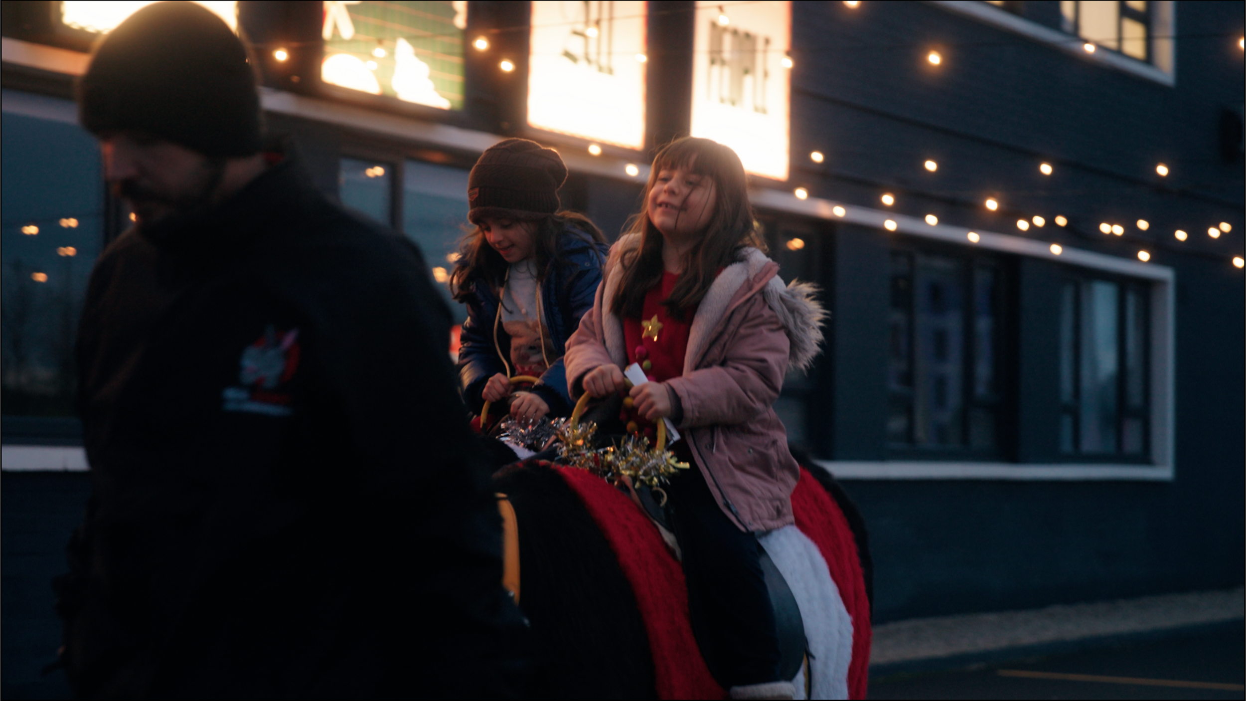 Two young girls riding a horse decorated for Christmas, accompanied by a man, during an outdoor evening event with string lights and a building in the background.
