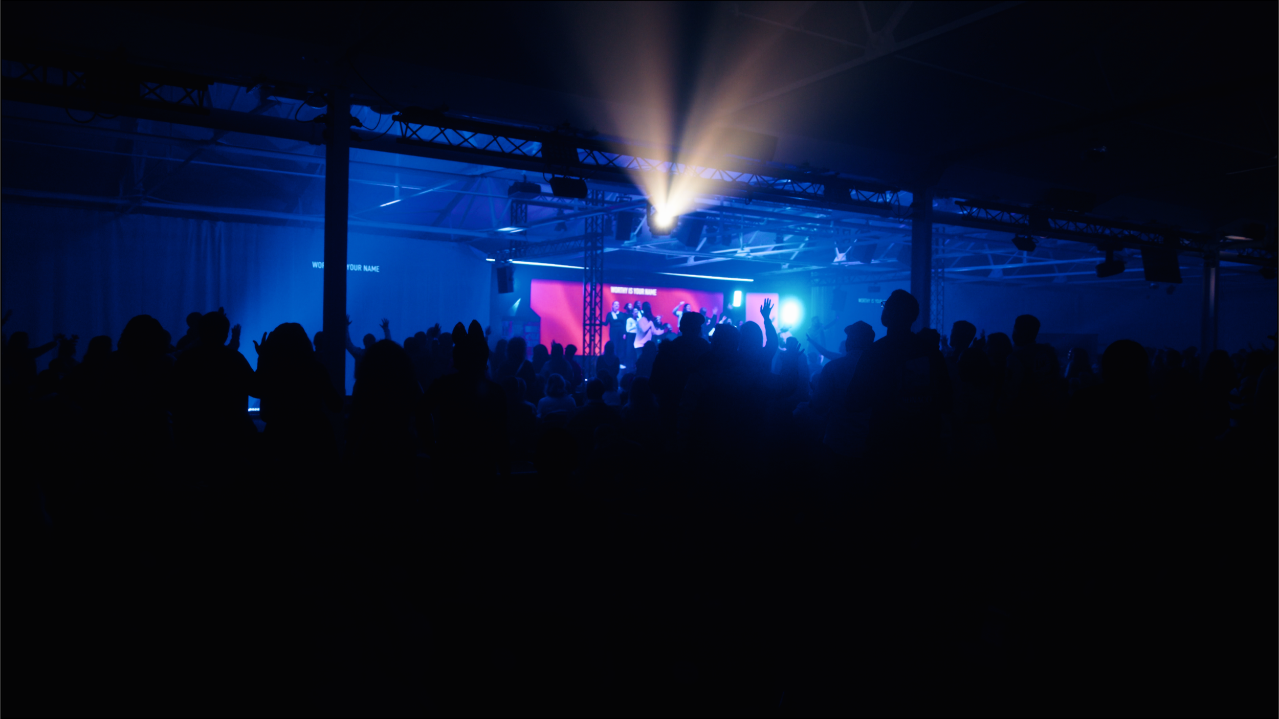 Crowd of people at concert or event with stage, blue lighting, and performers on screen