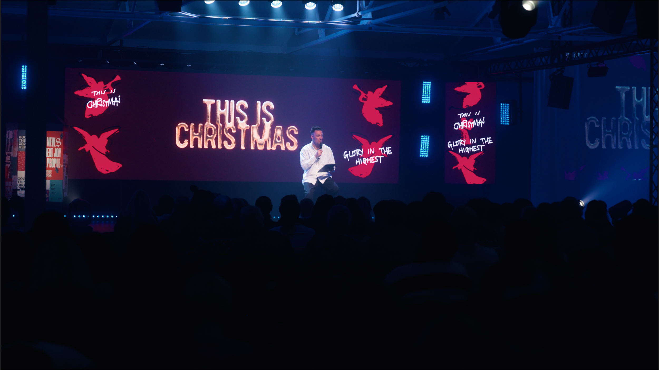 A man on stage speaking into a microphone in front of a large screen that says 'This is Christmas' with additional text 'Gluory in the highest'. The audience is seated in front of him, with stage lights illuminating the scene.