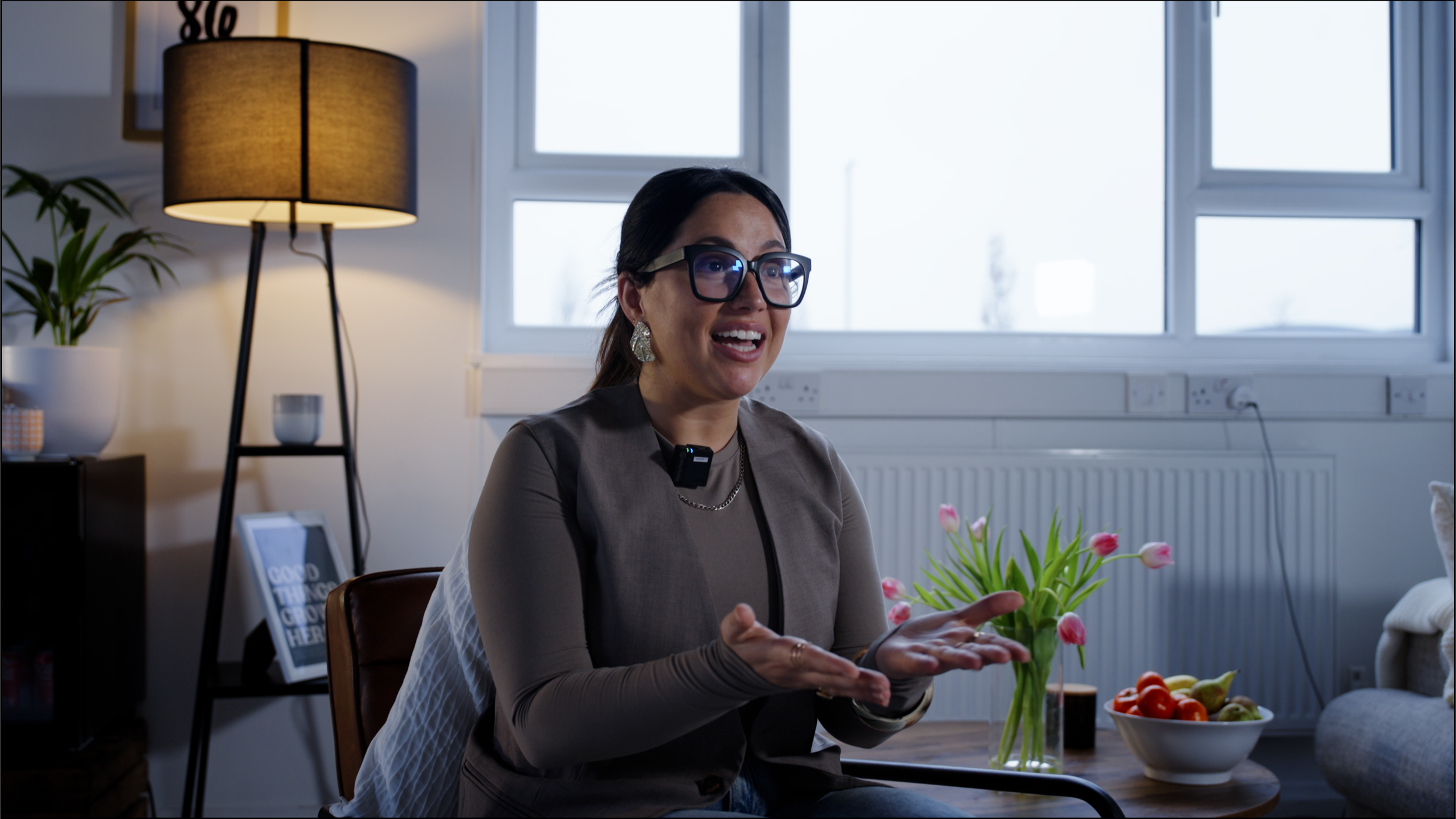 Woman with glasses and earrings talking and gesturing in a living room with a lamp, potted plant, framed quote, tulips, and a bowl of fruit on a table.
