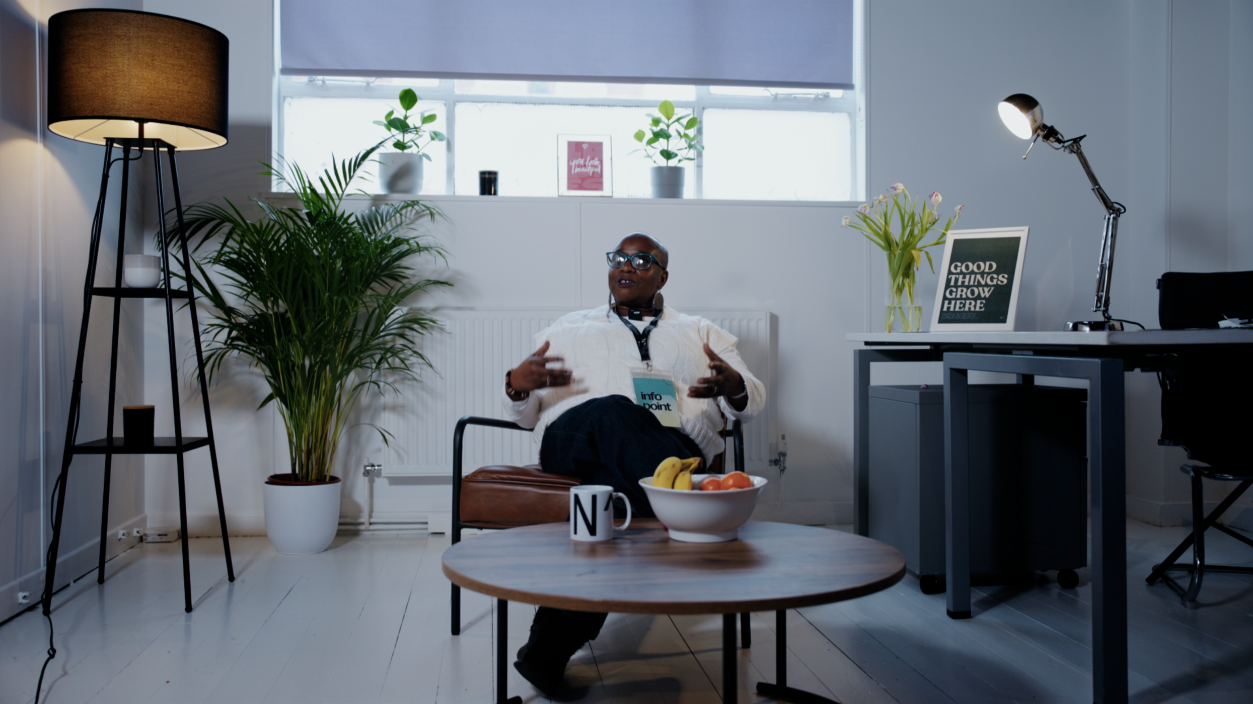 A woman with dark skin, short hair, glasses, white jacket, and layered necklaces sits in a modern office, gesturing as she talks. She is seated on a chair with a brown cushion. There is a large potted plant and a small table with a mug and a bowl of 