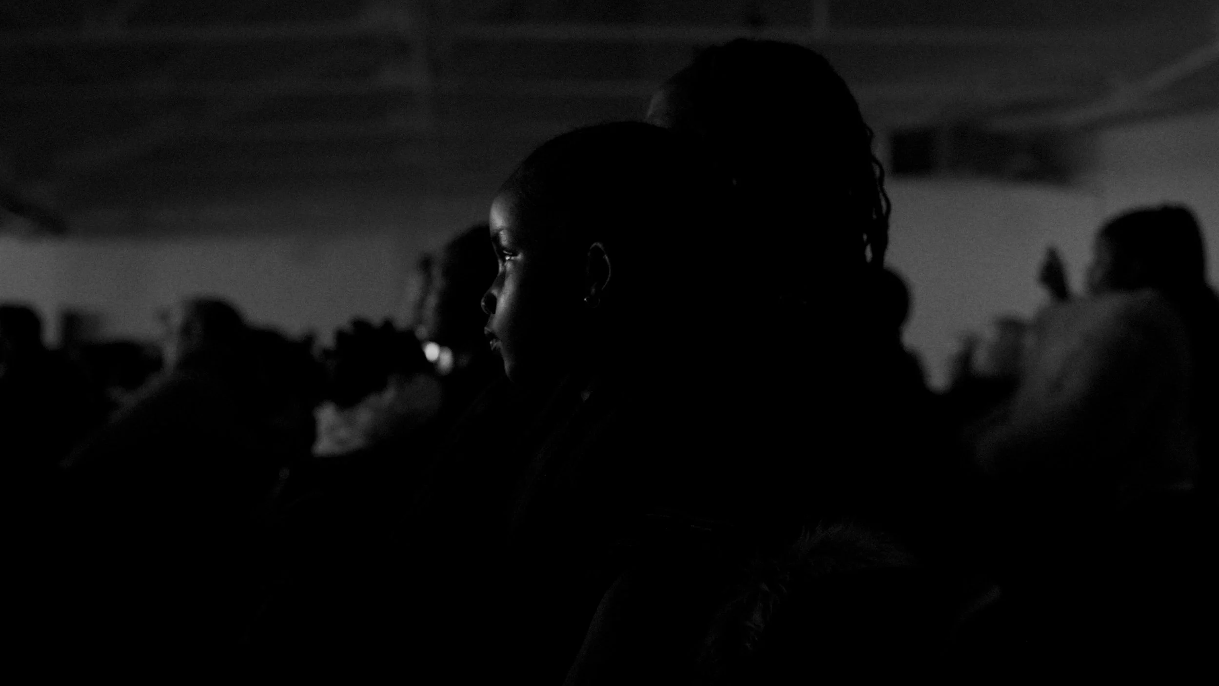 Side profile of a woman with earrings in a dark room filled with seated people, with the focus on her face illuminated softly.