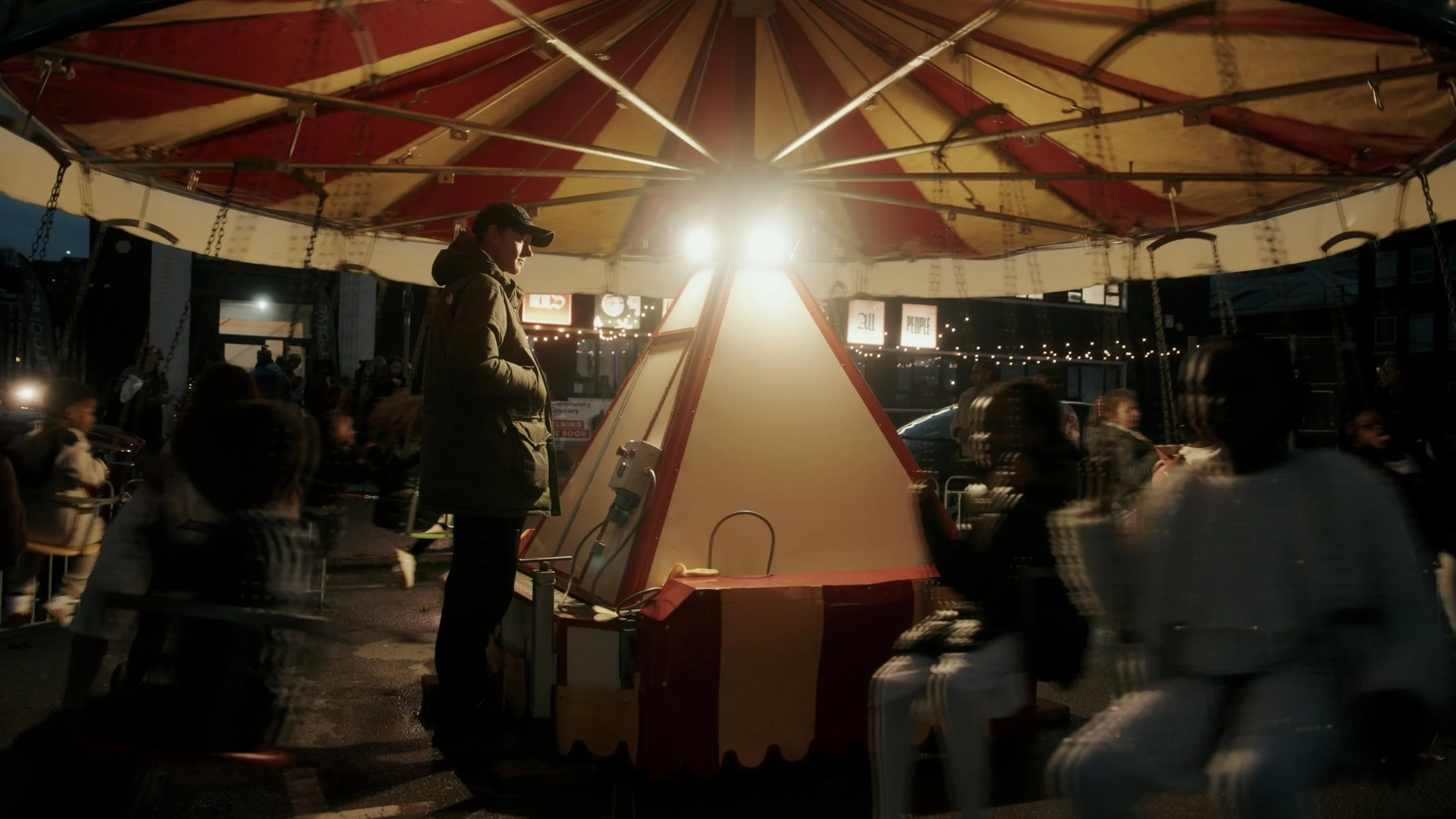 People riding a spinning amusement park ride at night with bright lights and a crowded background.