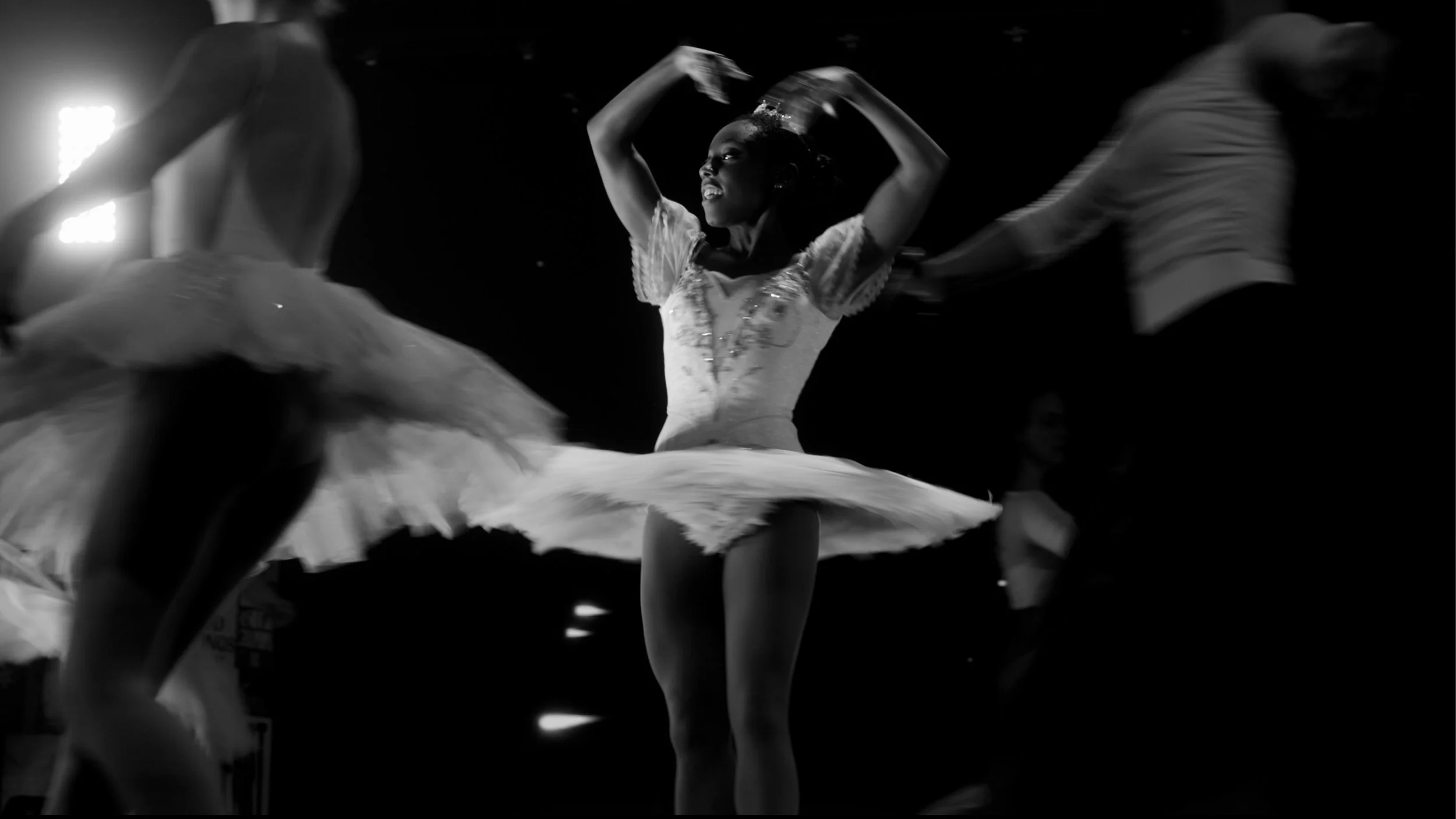 A ballerina in a tutu performing a dance on stage, with other dancers in the background.
