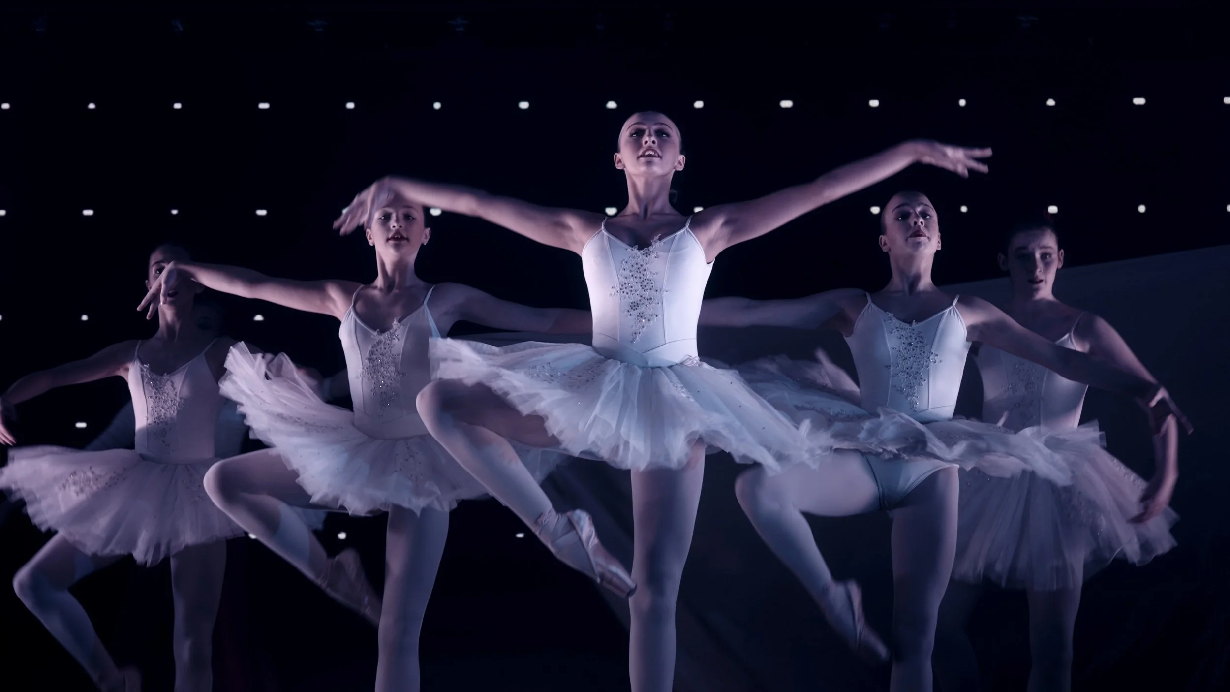 Ballet dancers performing on stage in white tutus and leotards with arms extended, under starry lighting
