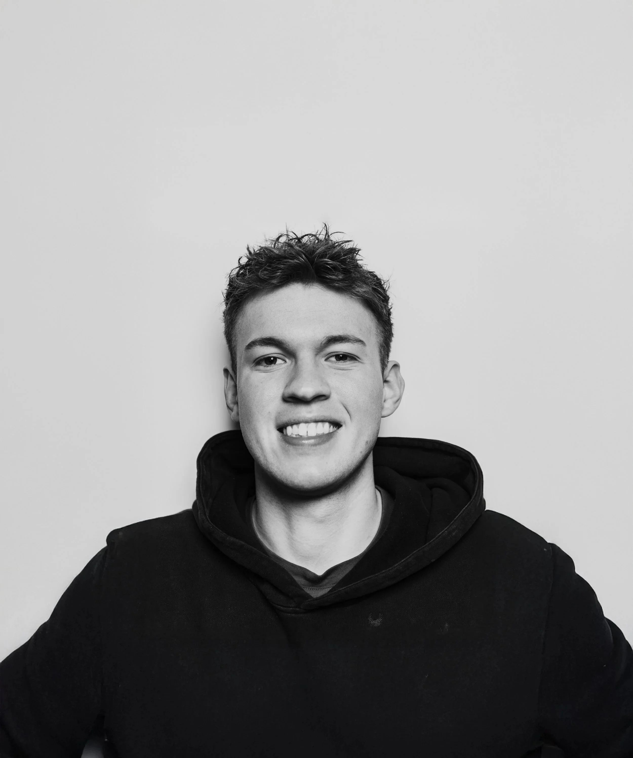 Black and white portrait of a smiling young man with short, curly hair wearing a dark hoodie, against a plain light background.