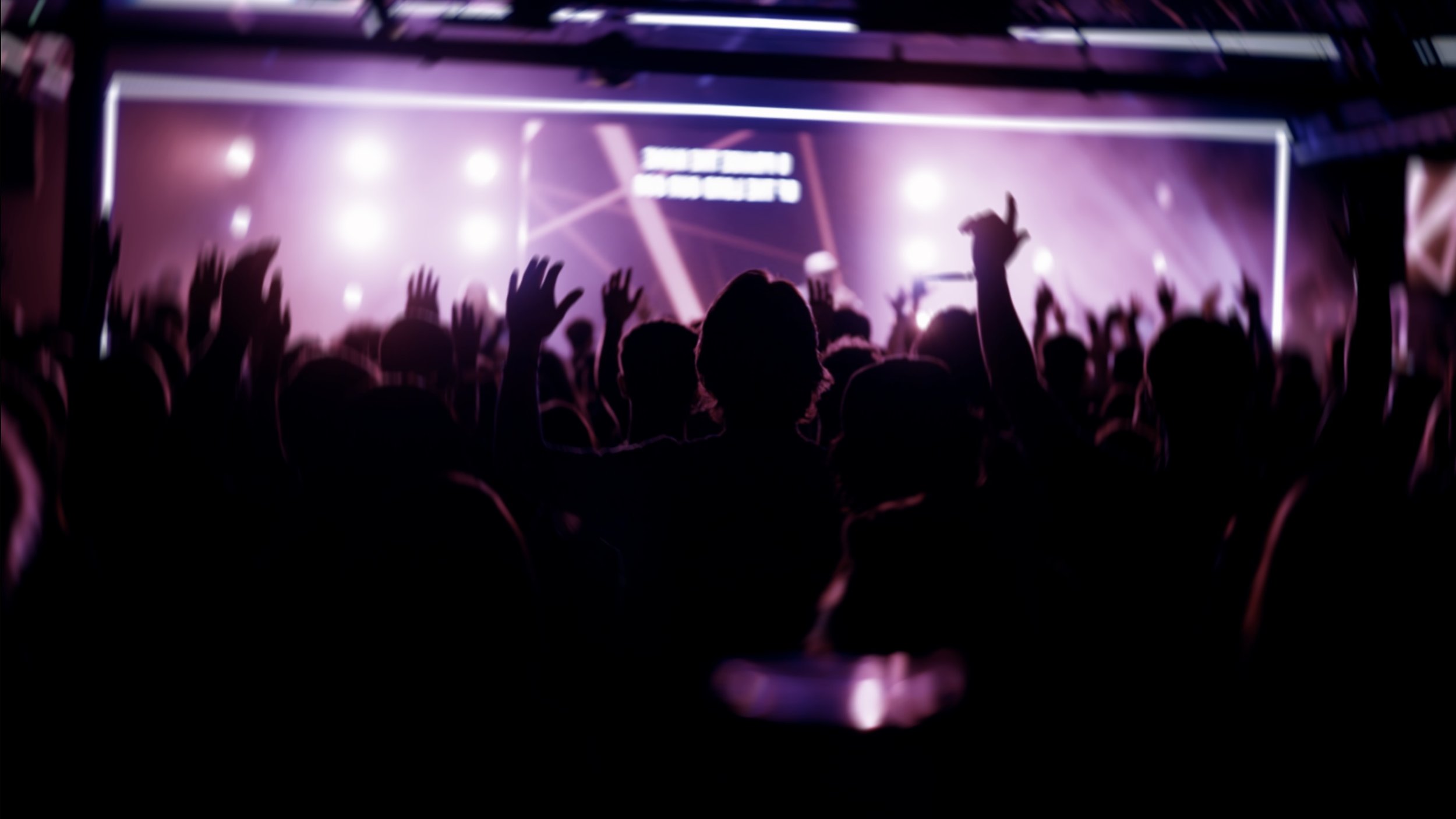 A crowd of people at a concert or music event with hands raised, facing a stage with purple and pink lighting and a screen with text in the background.