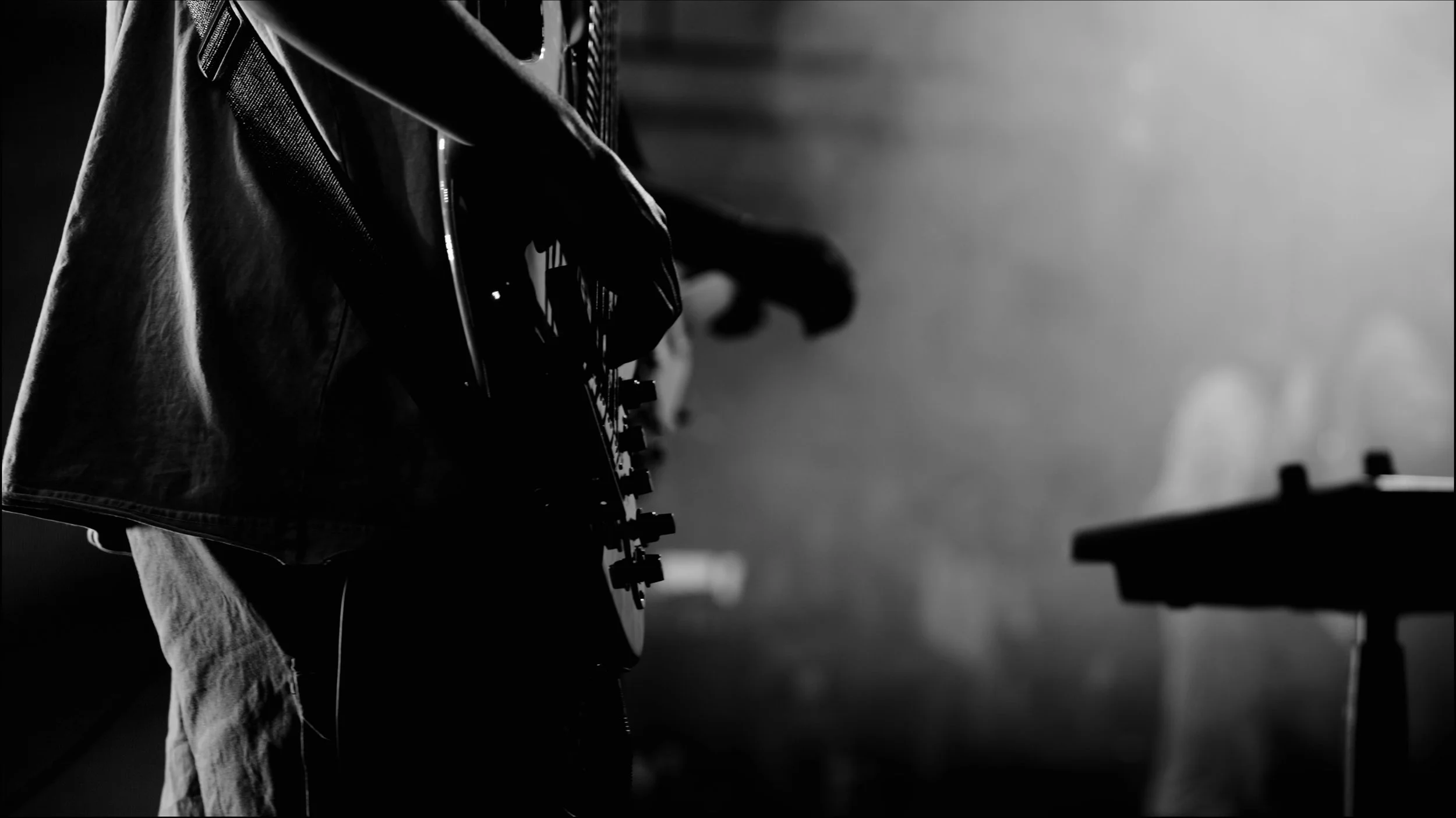 A person playing an electric guitar, seen in silhouette, in a dimly lit setting with a table and some bottles in the background.
