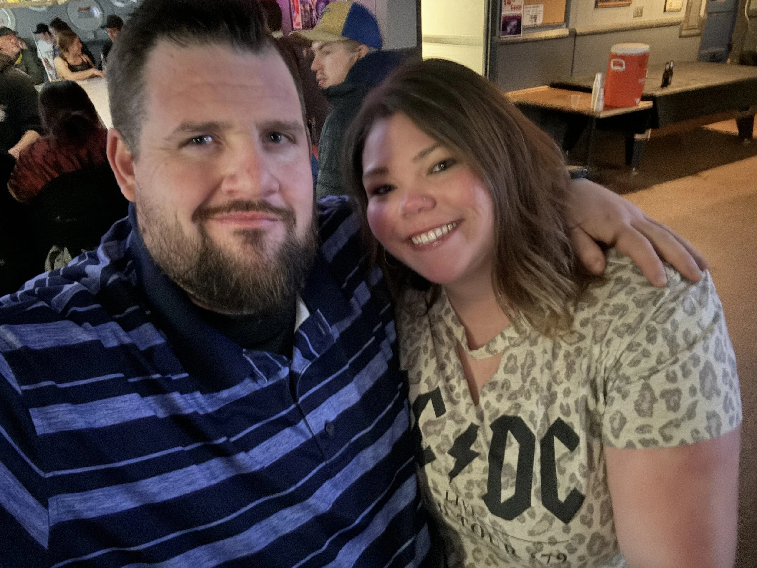 A man with a beard and a woman with a nose piercing smiling and taking a selfie in a crowded indoor setting.