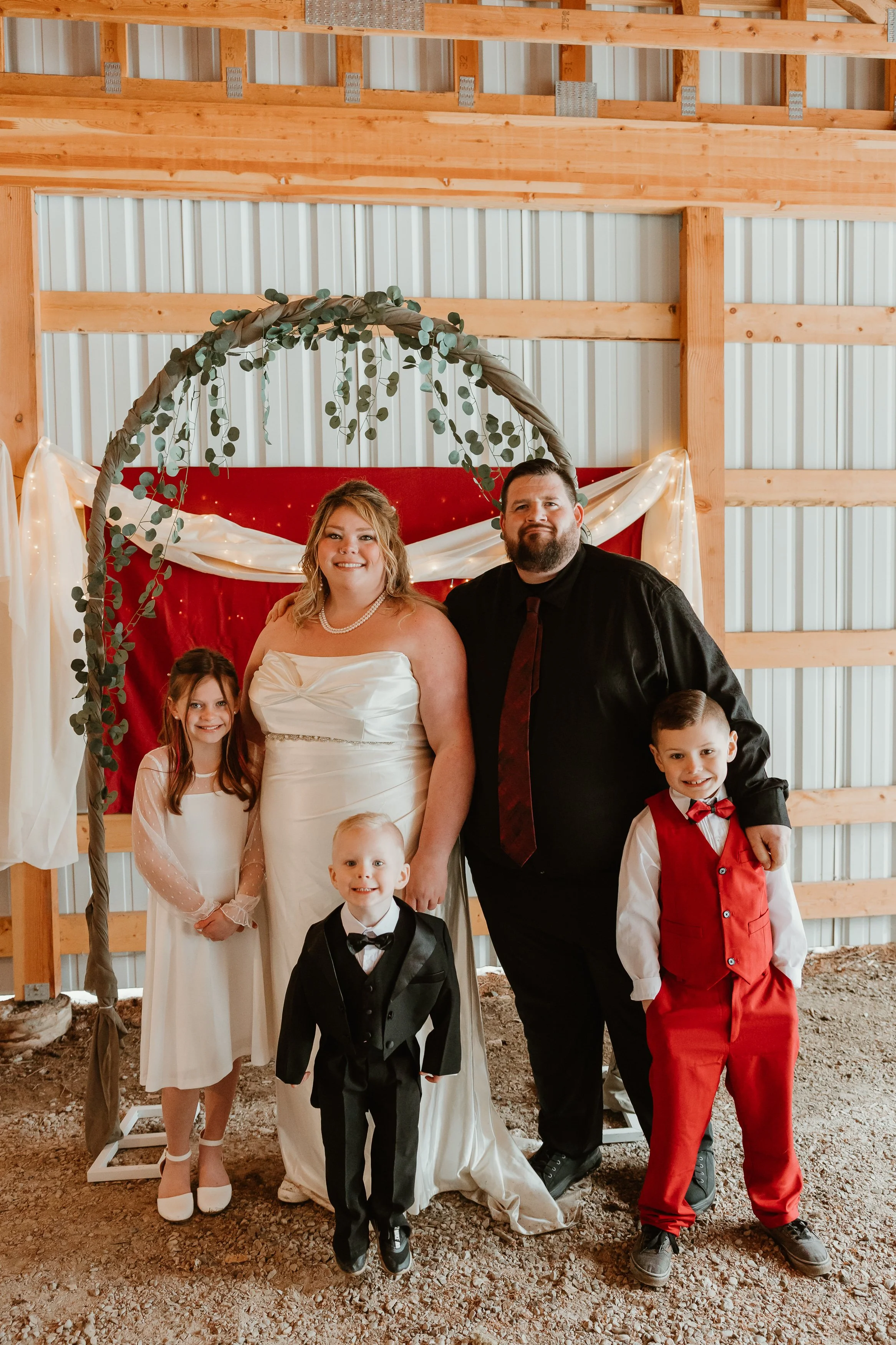 A family of five in wedding attire posing in front of a decorative backdrop inside a wooden barn, including a woman in a wedding dress, a man in black suit, a young girl in a white dress, and two young boys, one in a tuxedo and the other in a red suit.