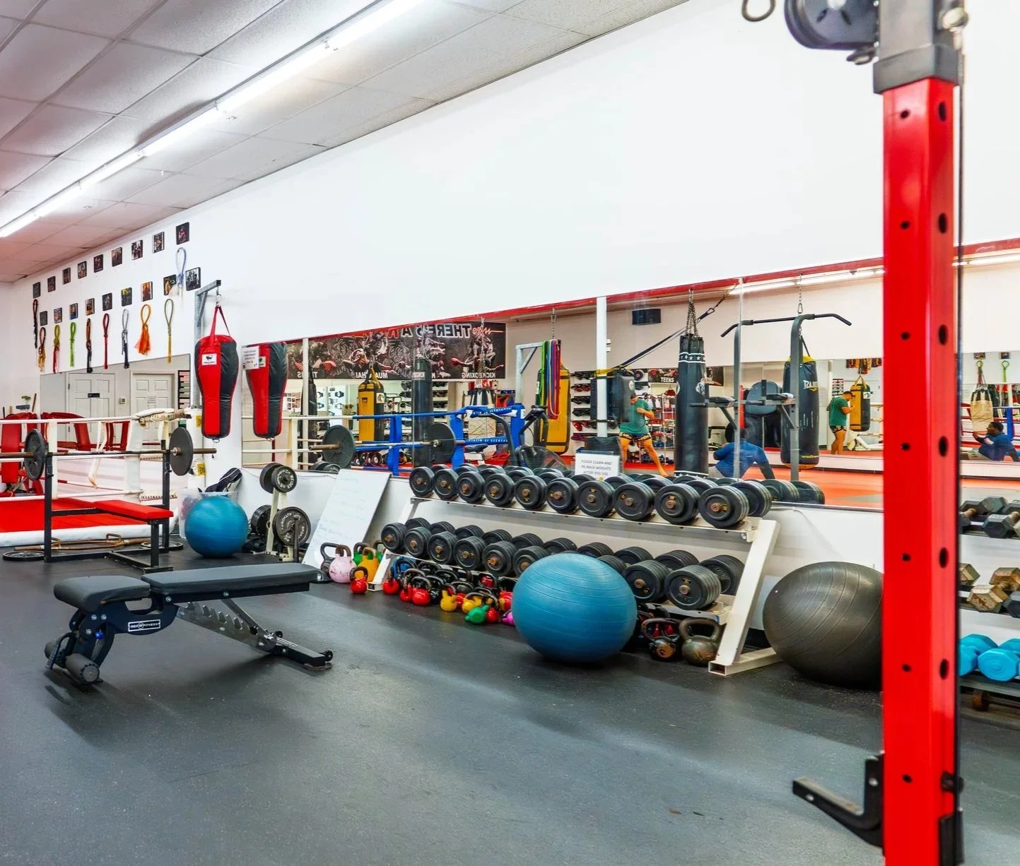A gym with various exercise equipment, including dumbbells, kettlebells, medicine balls, punching bags, and a weight bench, with a large mirror reflecting the workout area.