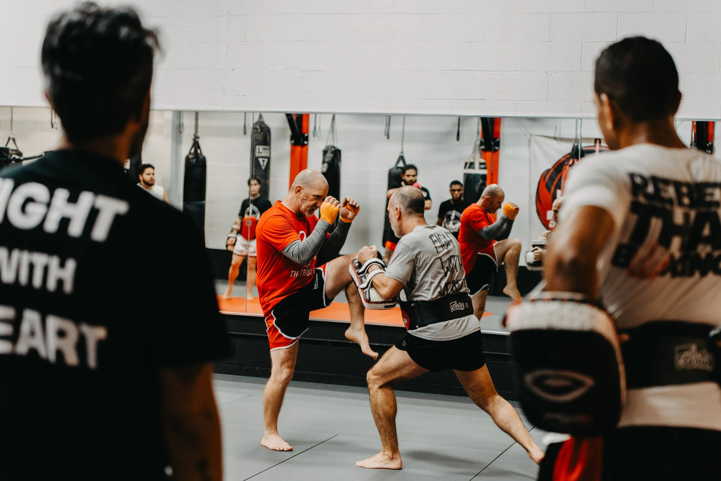 Two men practicing mixed martial arts in a gym with punching bags on the wall behind them, one holding a pad and the other in a fighting stance.